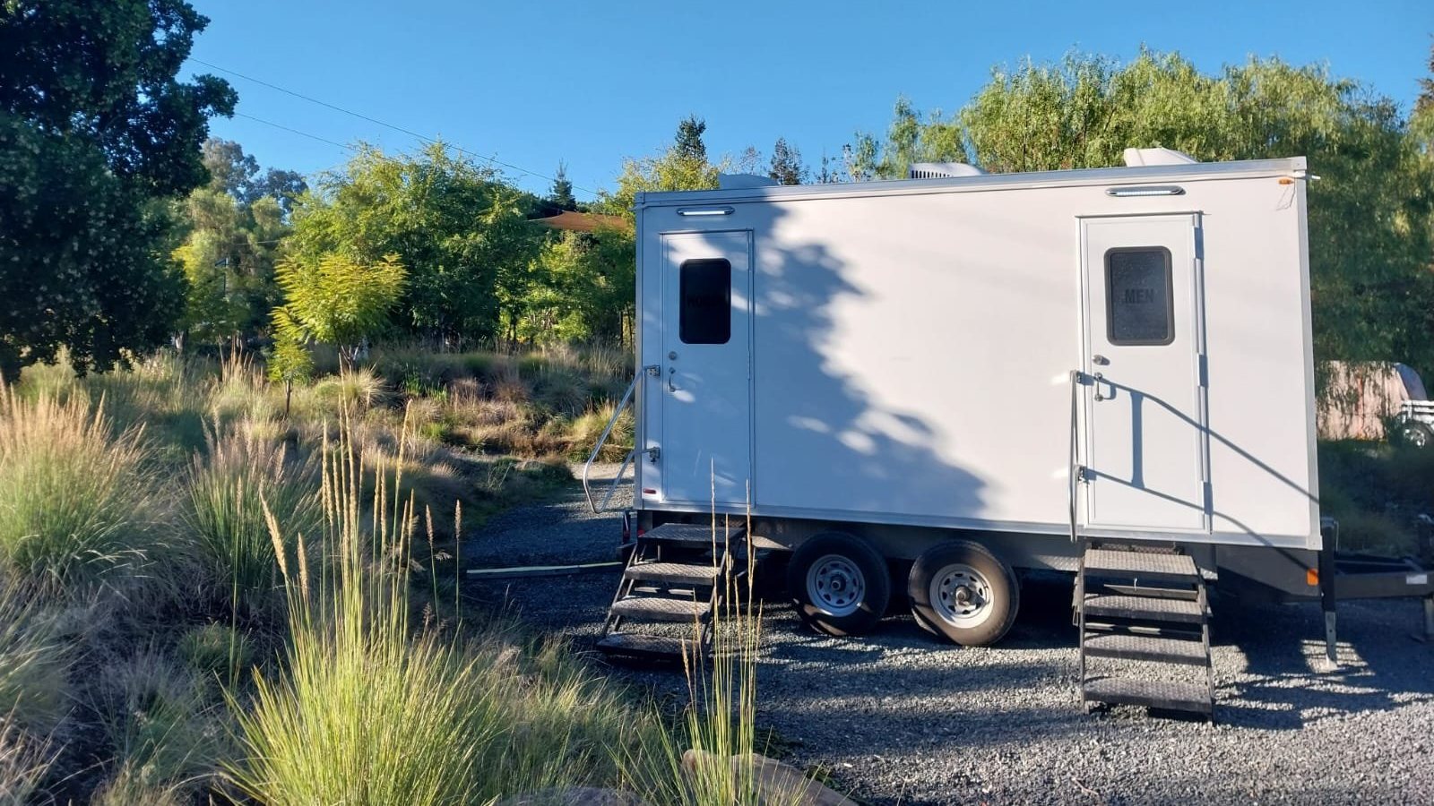 A Lavatory restroom trailer with separate men's and women's doors on a gravel lot beside tall ornamental grasses and trees