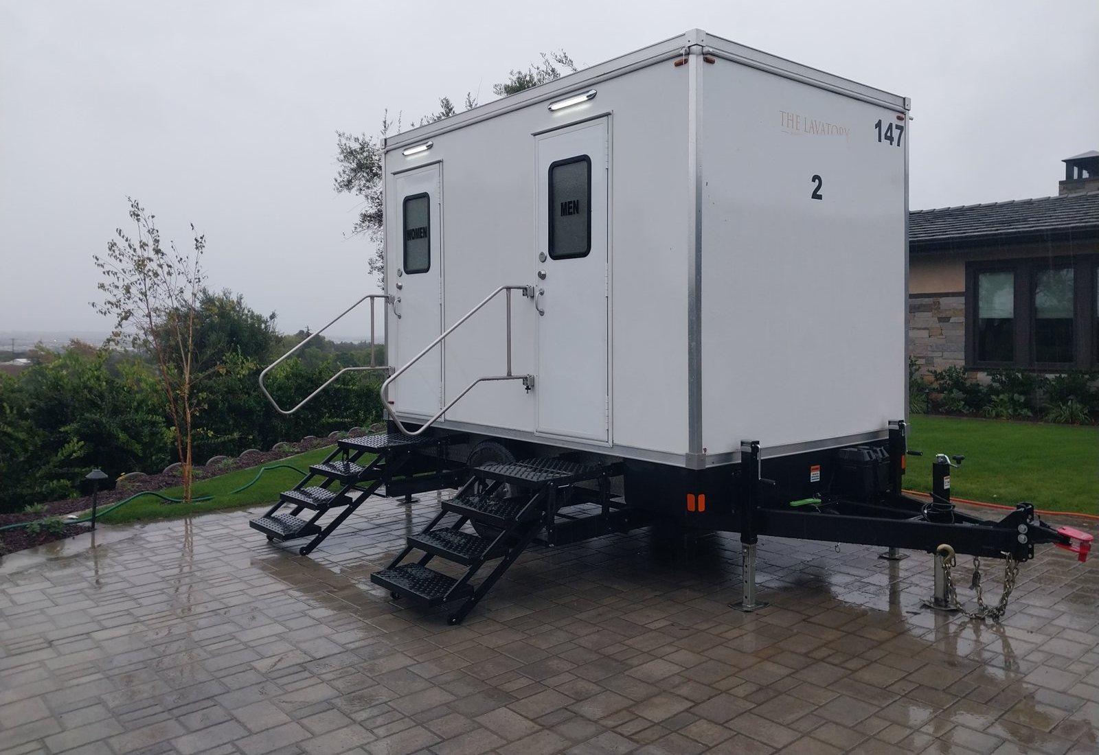 A Lavatory 2-Station restroom trailer on a wet paver patio beside a hilltop home on a rainy day