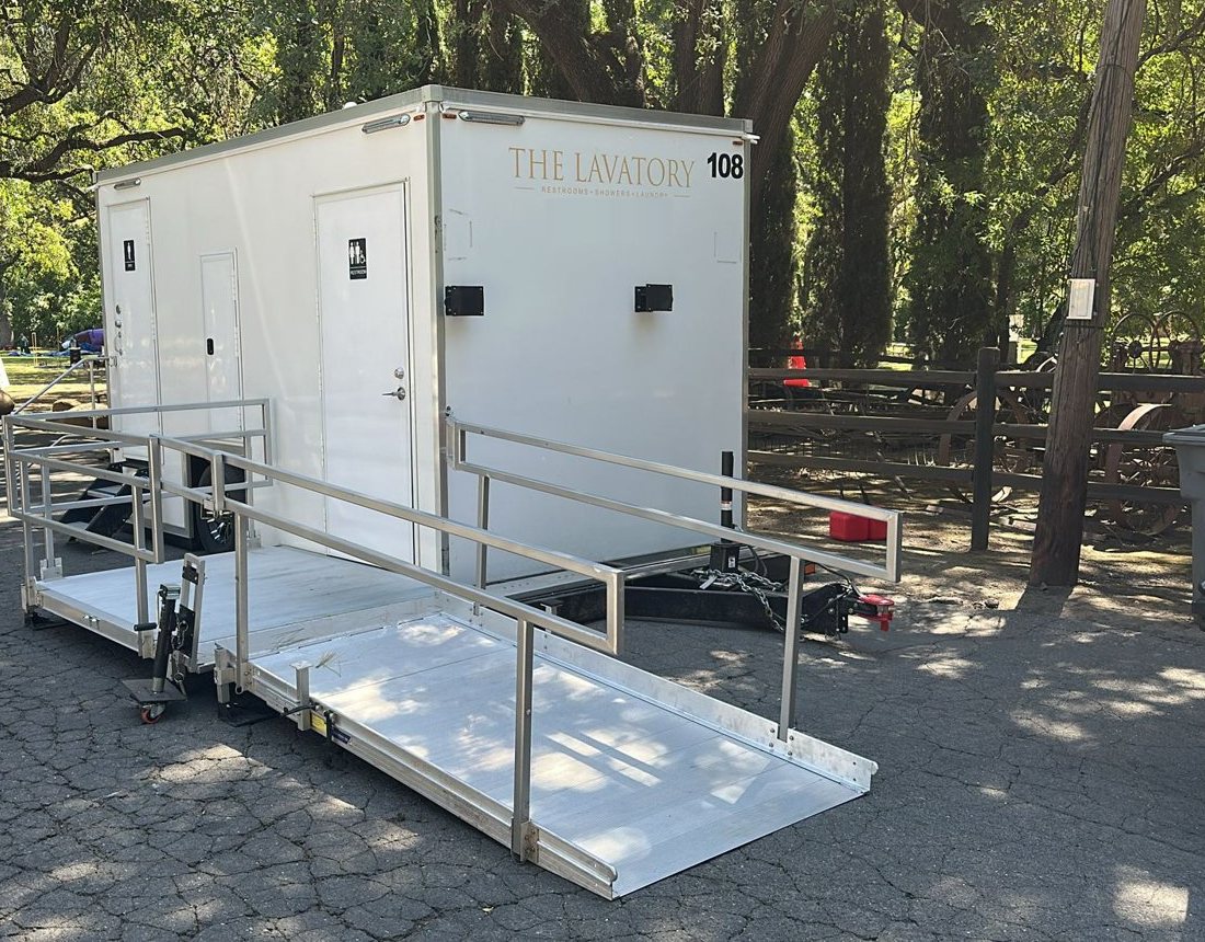 A Lavatory ADA restroom trailer with an aluminum ramp and handrails on a paved event lot under oak trees, with a vendor tent behind it