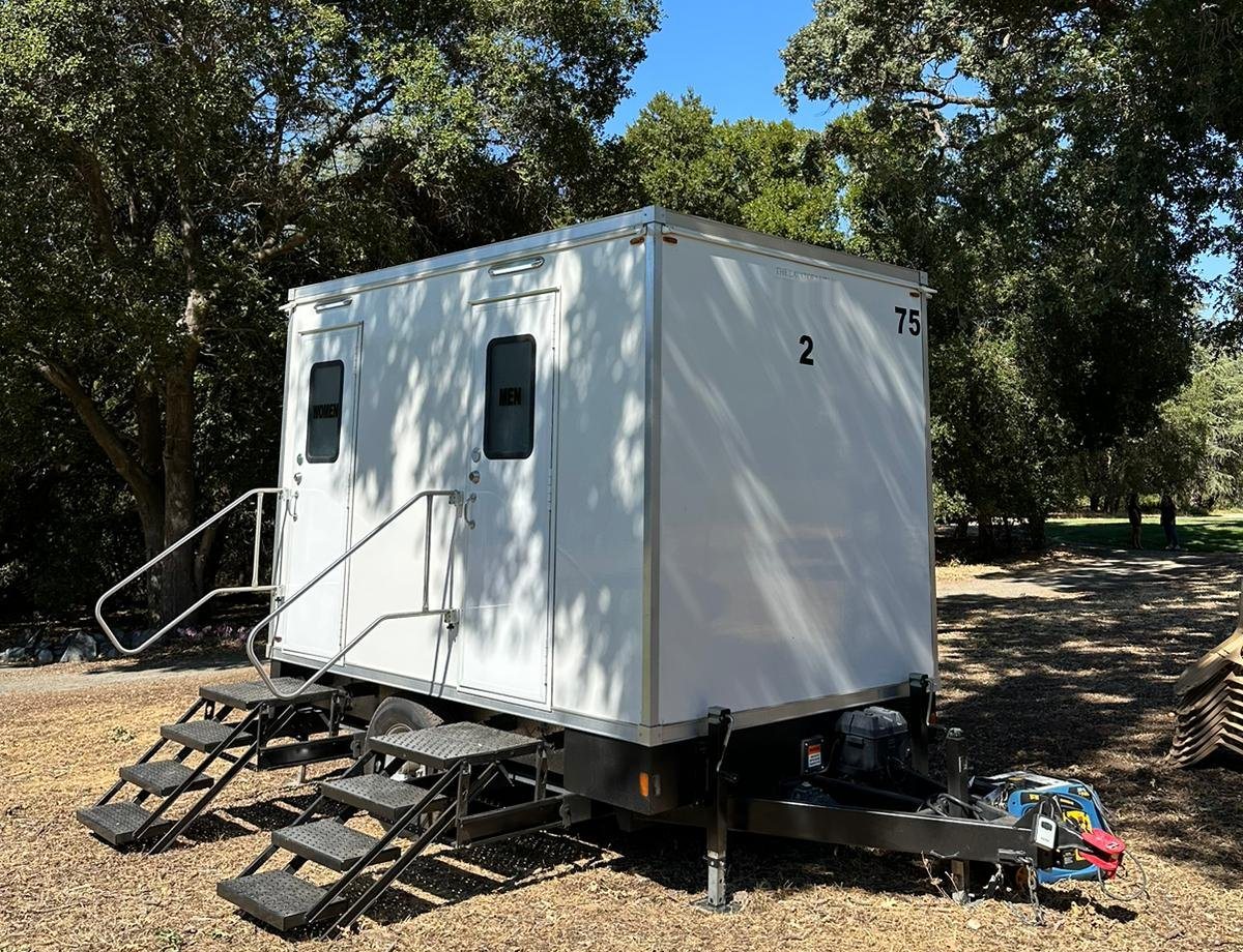 A Lavatory 2-Station restroom trailer with a portable generator under large oak trees at a park venue in Livermore