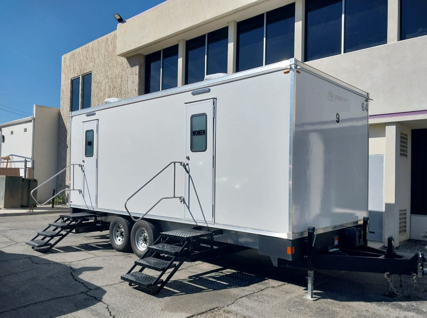 A Lavatory 9-Station restroom trailer with two sets of steel stairs parked beside a glass-front office building