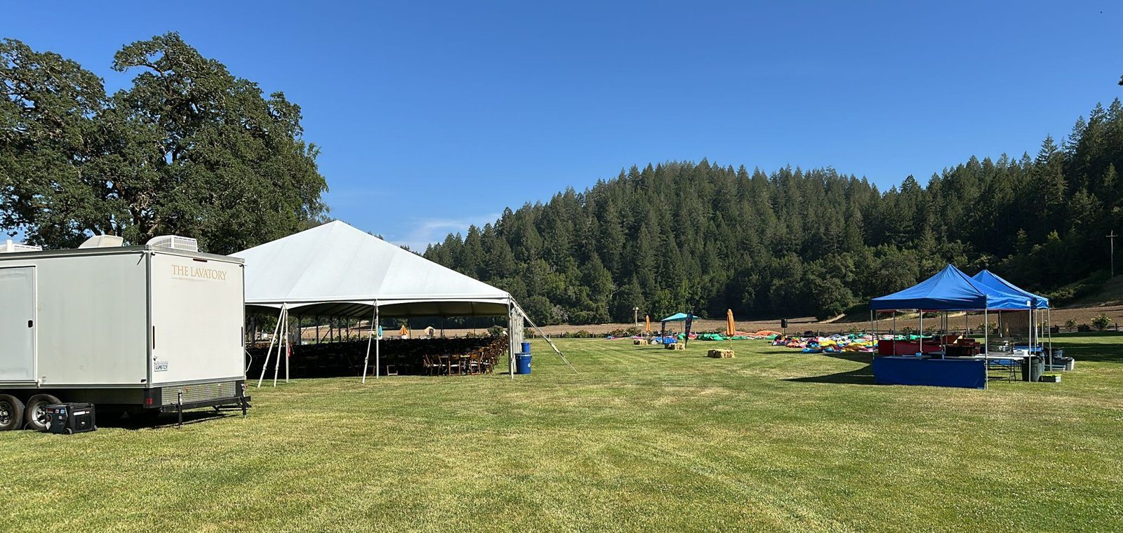 A Lavatory restroom trailer and a quiet generator beside a white reception tent on a mowed lawn, with vendor canopies and a wooded hillside behind
