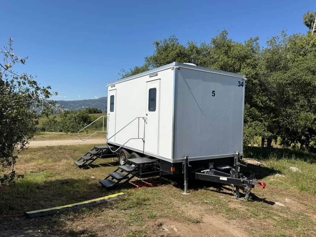A Lavatory 5-Station luxury restroom trailer set on dry grass beside oak trees, with hills in the distance