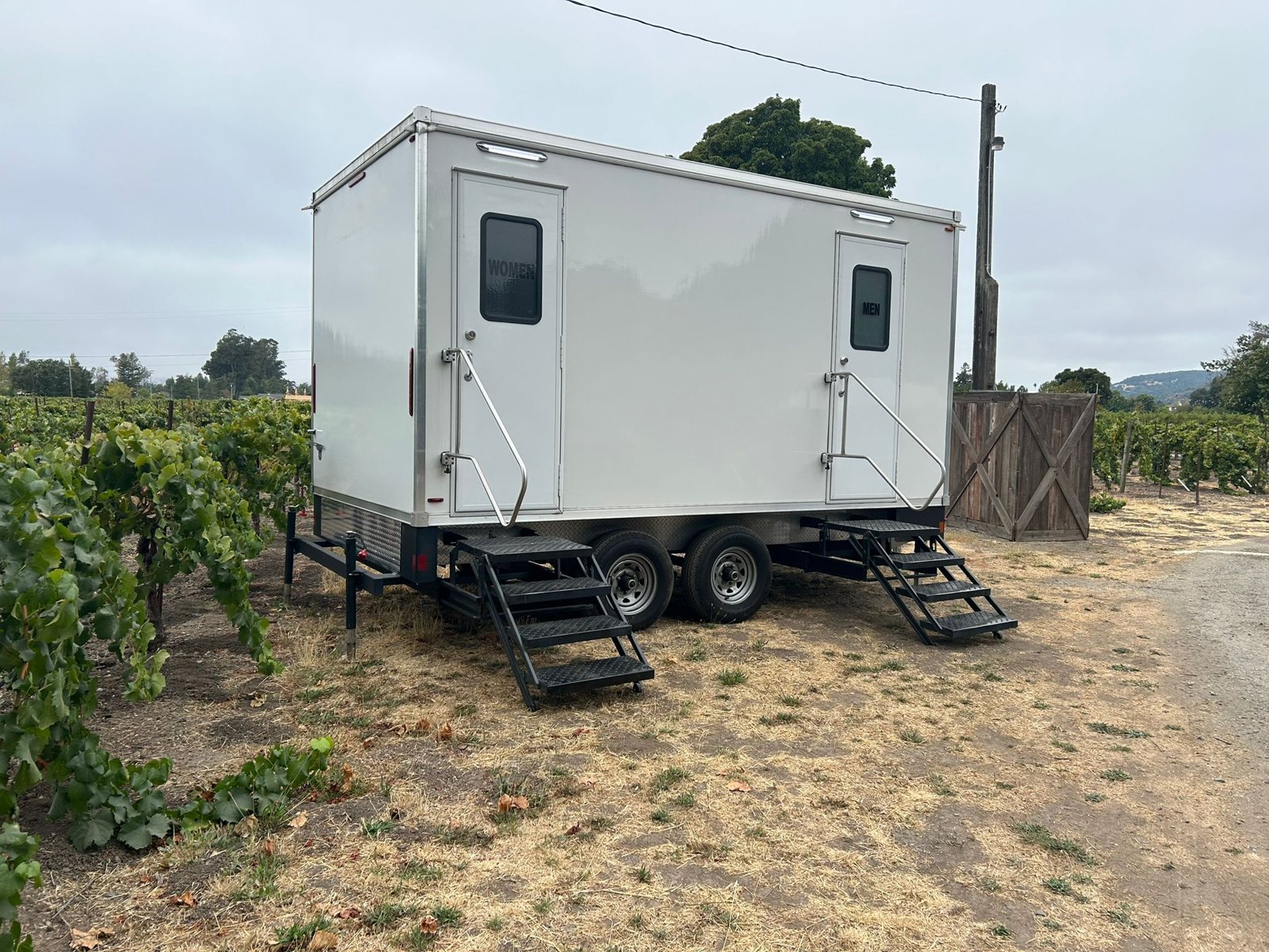 A Lavatory 2-Station luxury restroom trailer parked on a dirt lane beside grapevines, with a wooden gate and hills in the distance