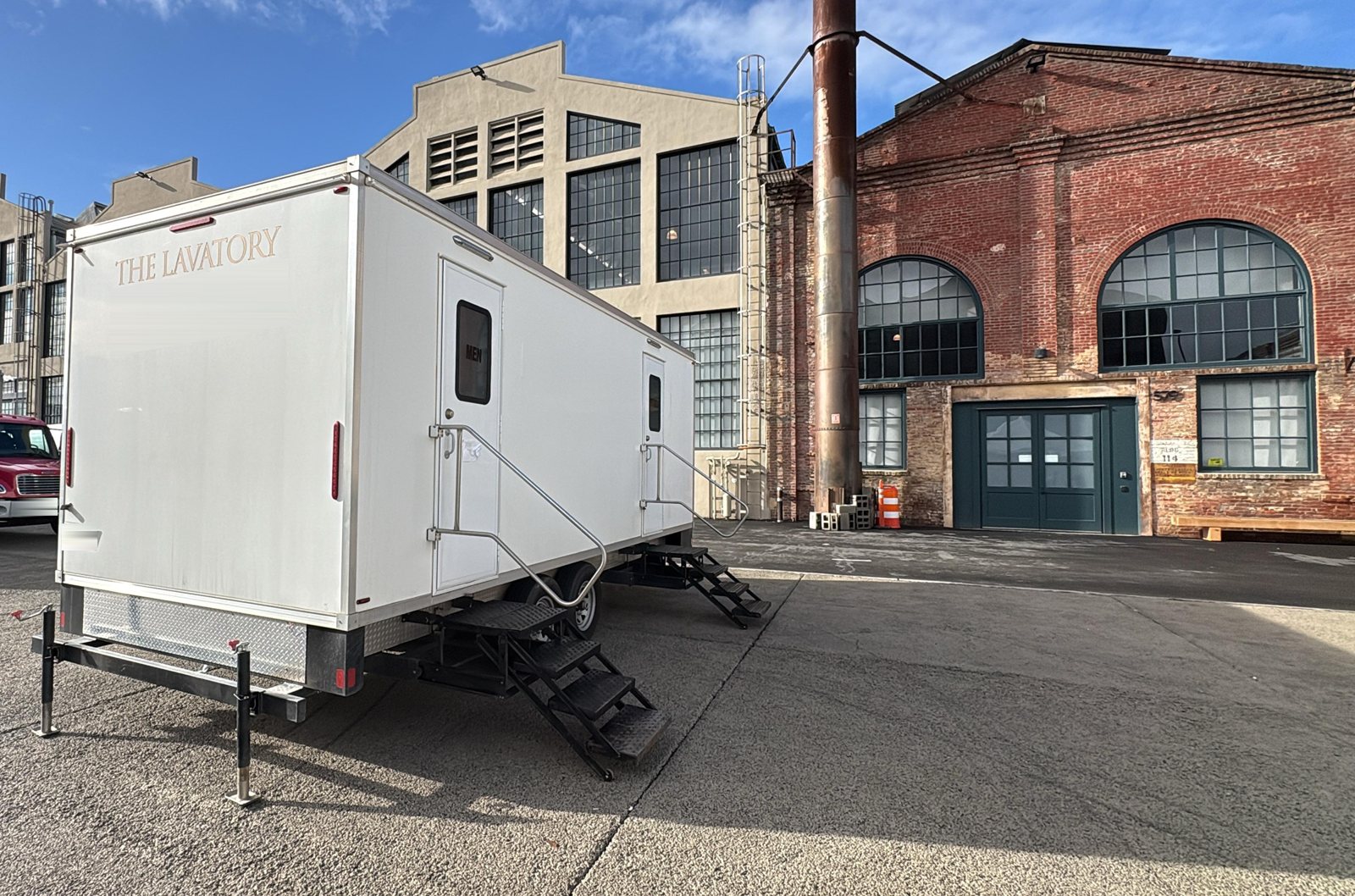 A Lavatory 9-Station restroom trailer with its steps down on an industrial lot between an old brick warehouse with arched windows and a tall factory building