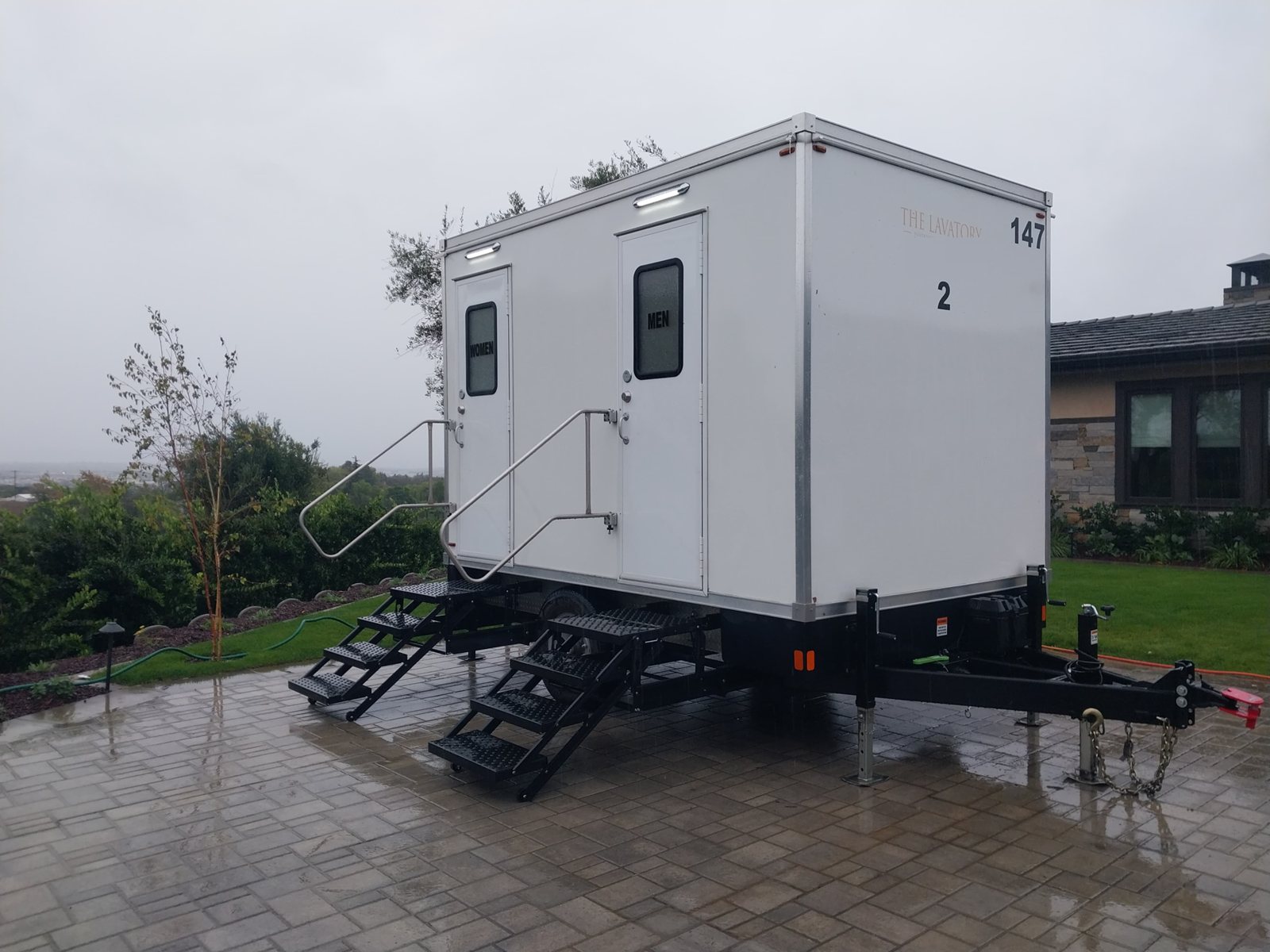 A Lavatory 2-Station restroom trailer on a wet paver patio beside a hillside home on a gray, rainy day