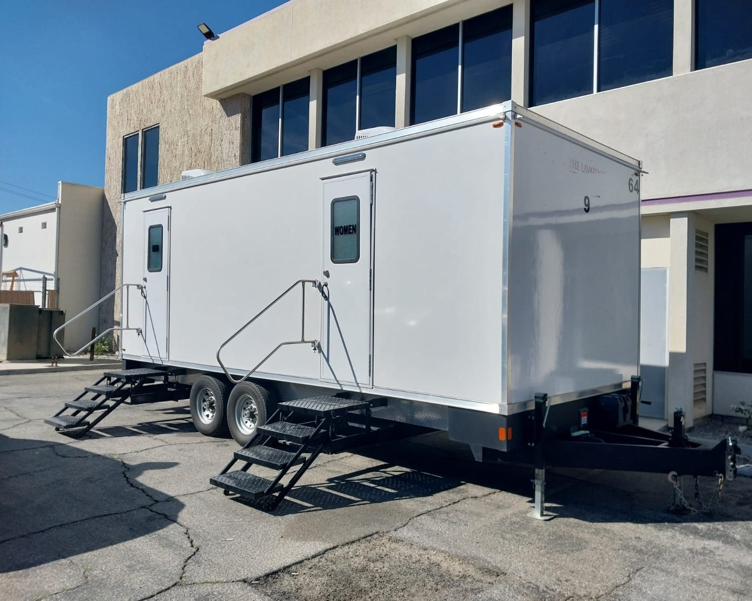 A Lavatory 9-Station restroom trailer with women's and men's doors parked beside a two-story office building