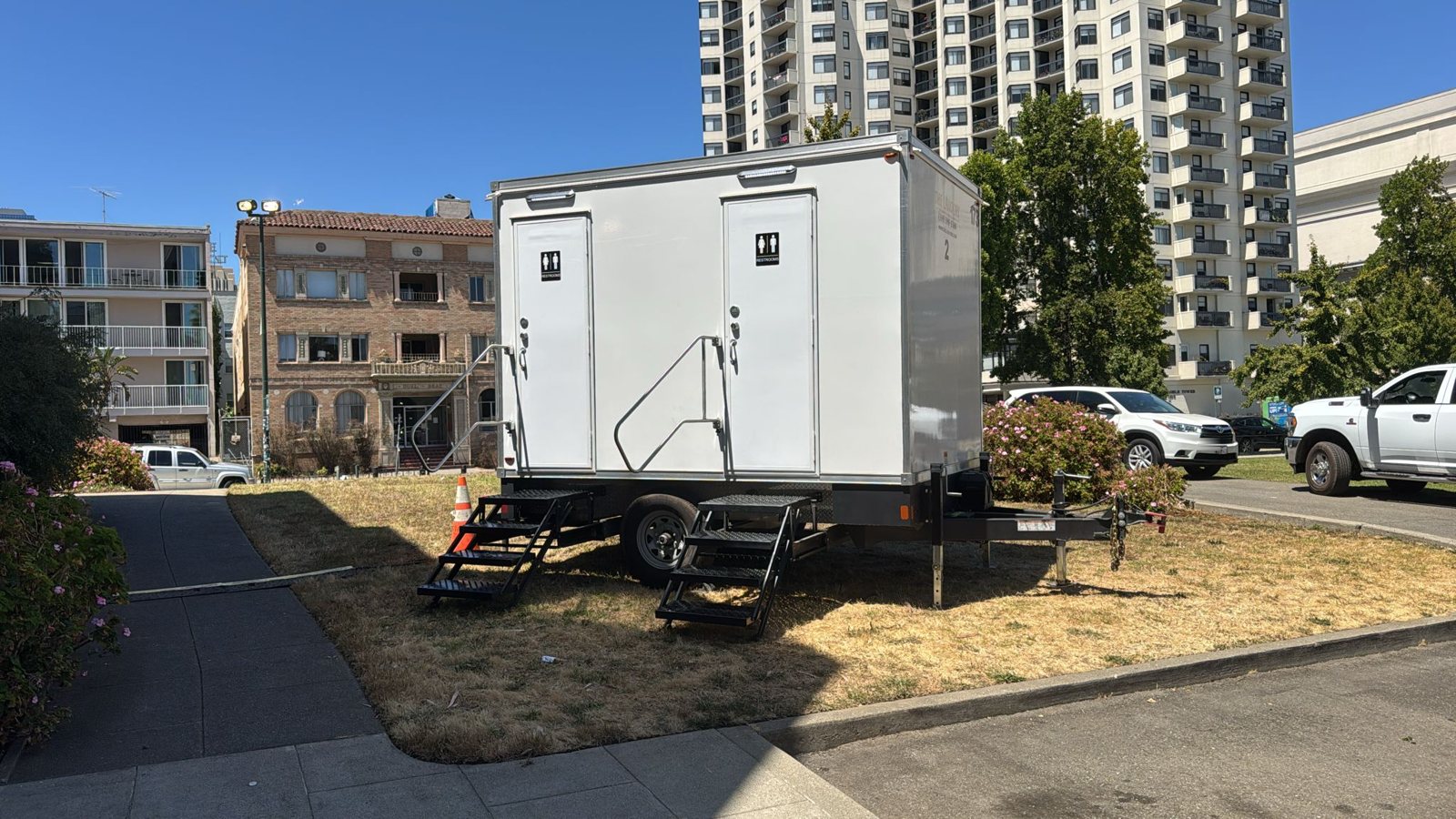 A Lavatory 2-Station restroom trailer on a grass city lot between apartment buildings and a high-rise tower