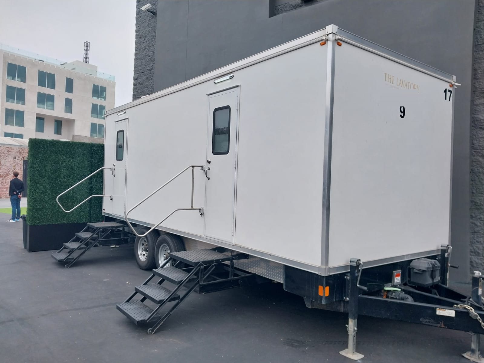 A Lavatory 9-Station restroom trailer set beside a boxwood hedge wall at an urban event venue on an overcast day