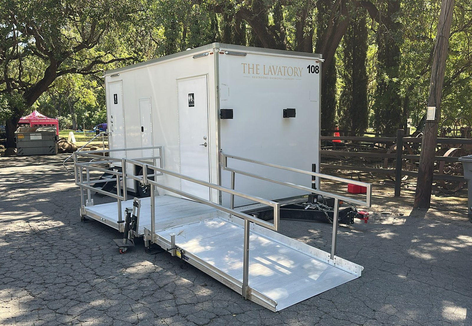 A Lavatory ADA restroom trailer with a railed aluminum ramp on a shaded park lot near vendor tents