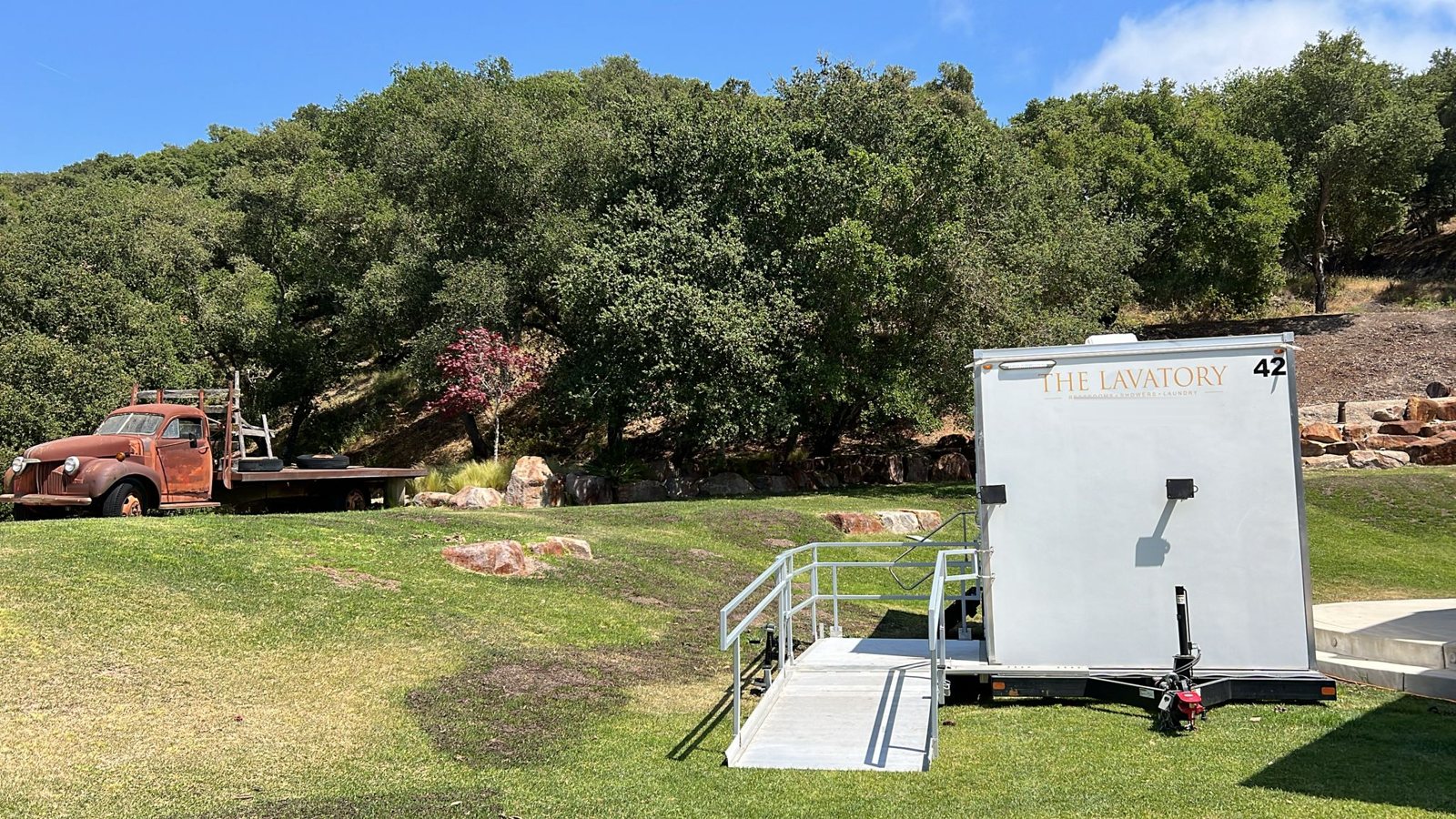 A Lavatory ADA restroom trailer with its ramp on a green lawn at an oak-covered ranch venue with a vintage flatbed truck