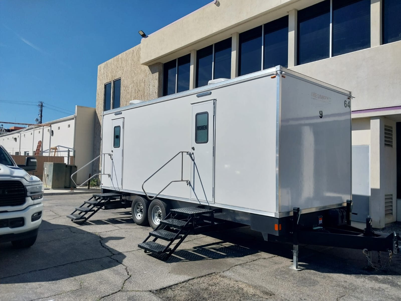 A Lavatory 9-Station restroom trailer parked beside a two-story office building with a pickup truck nearby