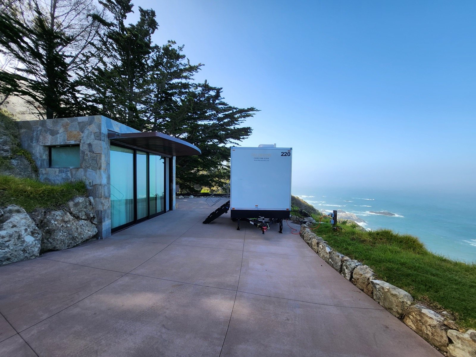 A Lavatory restroom trailer on a paved terrace beside a modern stone house, with the ocean below