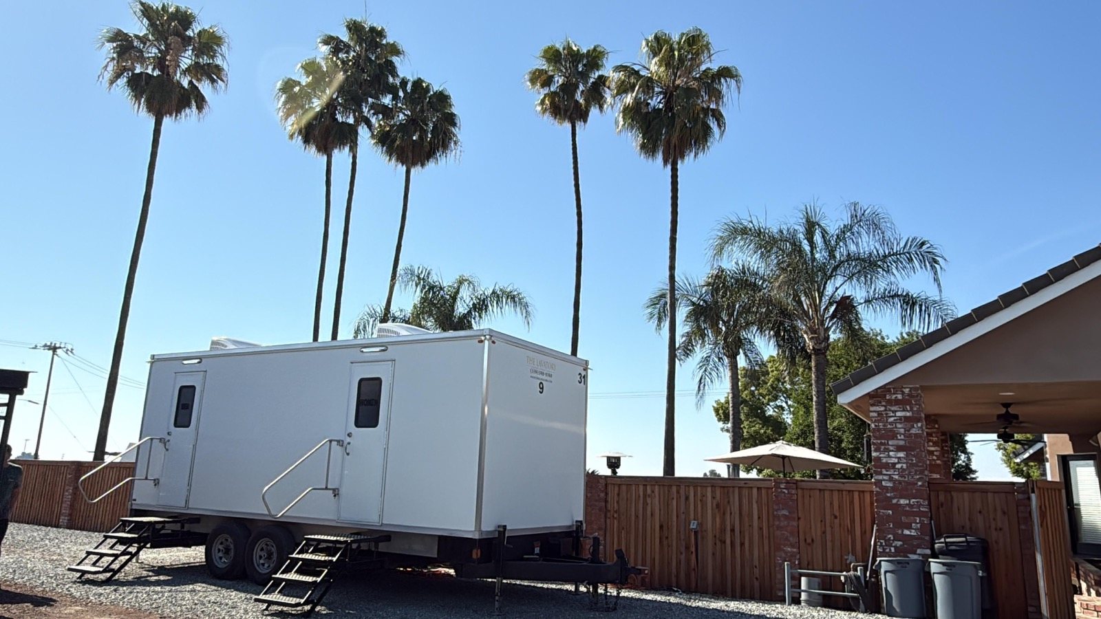 A Lavatory 9-Station restroom trailer on a gravel pad beside a wood fence and covered patio, with tall palm trees above