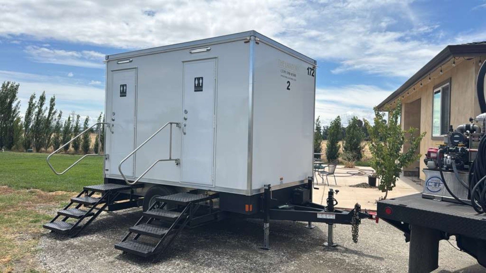 A Lavatory 2-Station restroom trailer on gravel beside a stucco building, with a service truck parked next to it