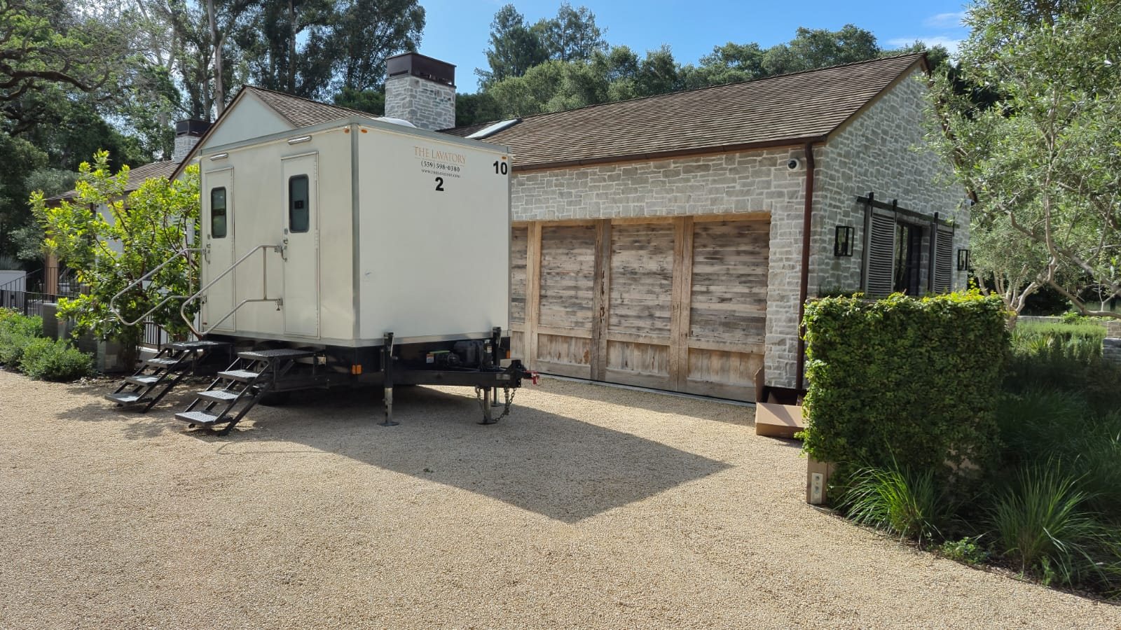 A Lavatory 2-Station restroom trailer on a gravel drive beside a stone house with wooden garage doors