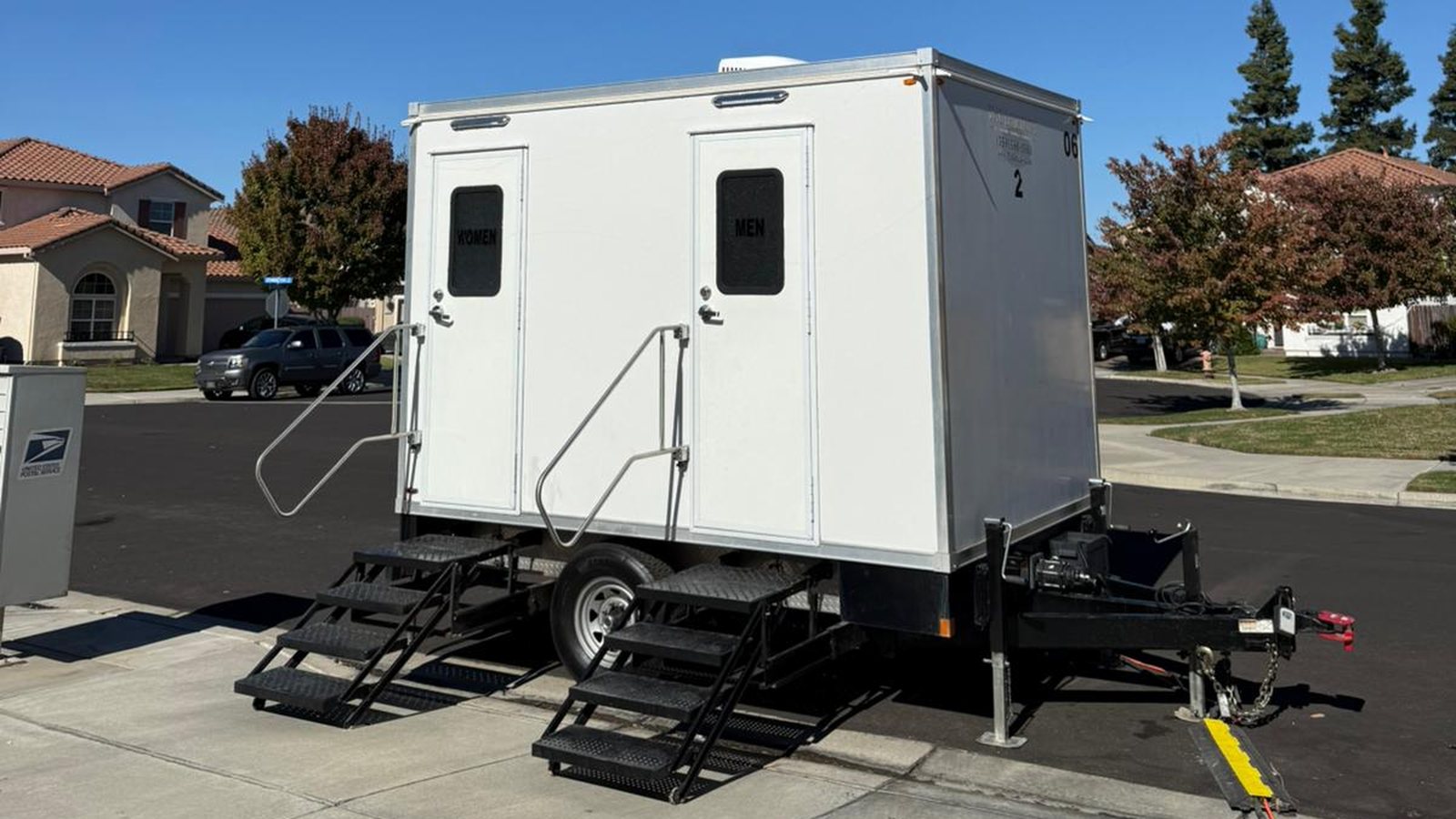 A Lavatory 2-Station luxury restroom trailer with steel stairs and handrails parked at the curb of a residential street in Turlock
