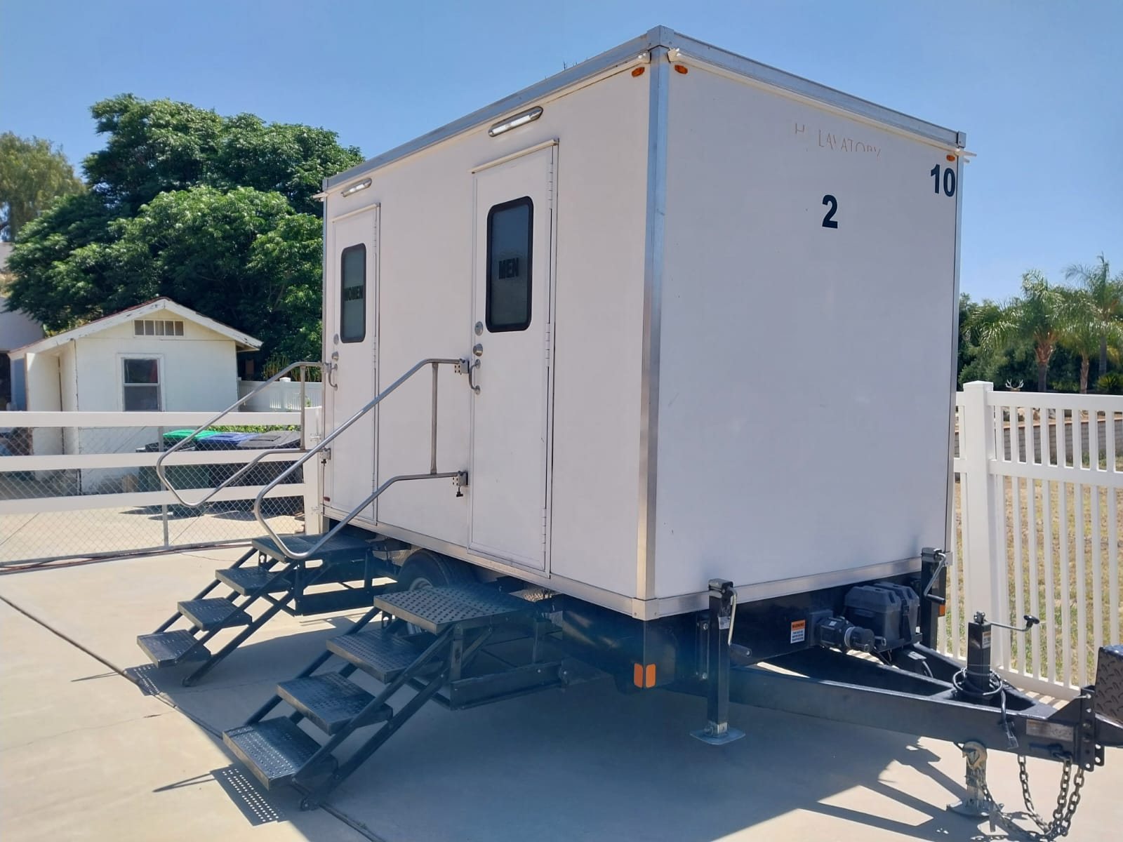 A Lavatory 2-Station restroom trailer parked on a concrete driveway beside a white vinyl fence at a private home