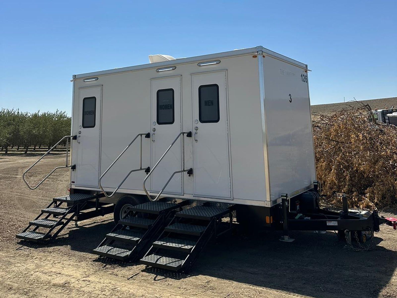 A Lavatory 3-Station restroom trailer with three doors and steel stairs on a dirt pad at the edge of an almond orchard