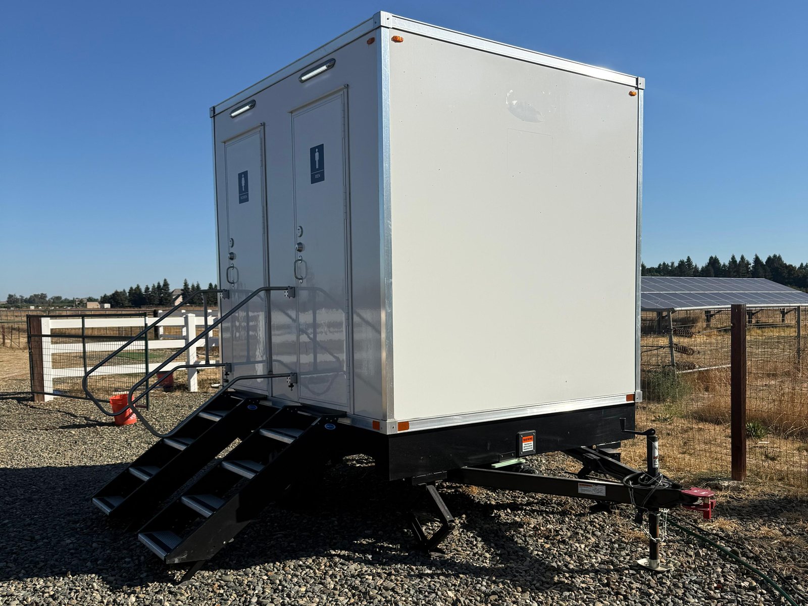 A Lavatory 2-Station restroom trailer on a gravel pad at a rural ranch with a white rail fence and open fields