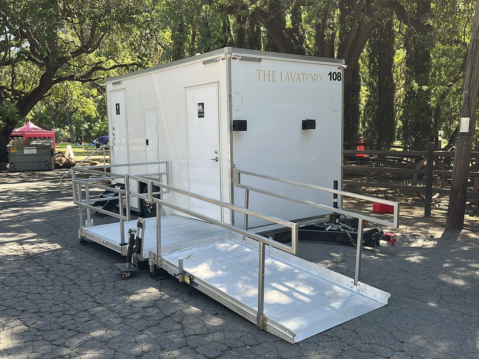 A Lavatory ADA restroom trailer with an aluminum ramp and handrails set up on a paved lot under shade trees at an outdoor event