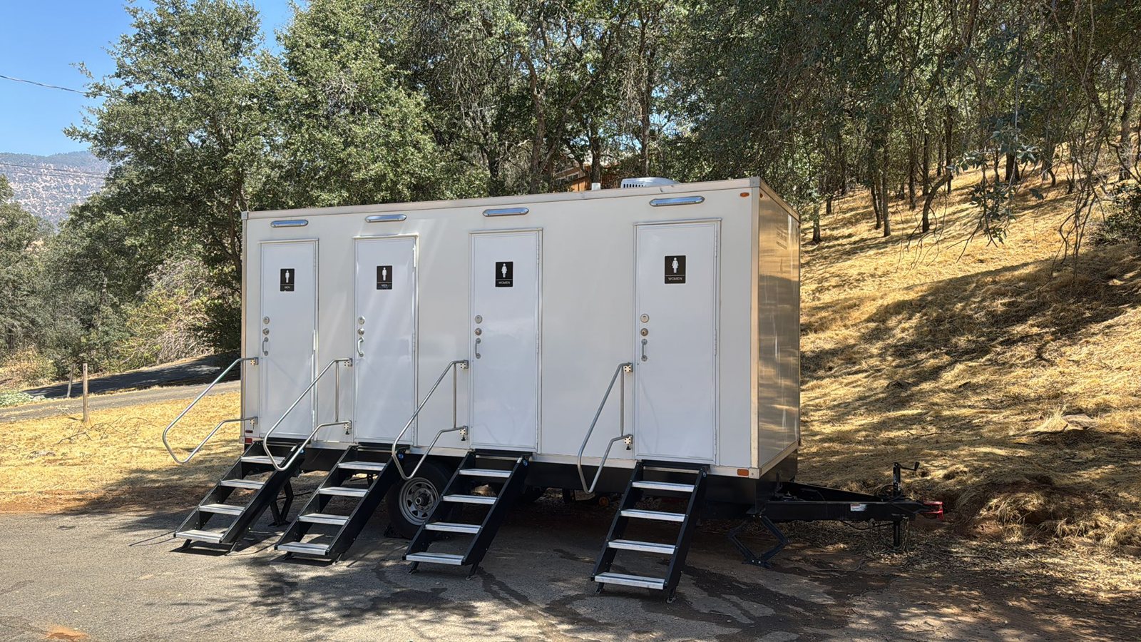 A Lavatory four-door luxury restroom trailer with steel stairs and handrails parked under oak trees beside a dry grass hillside