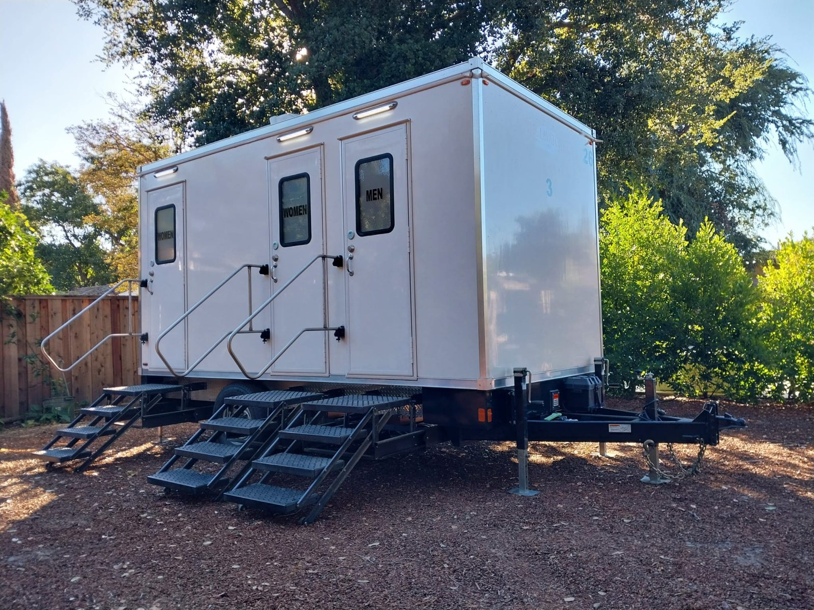 A Lavatory 3-Station restroom trailer on a bark-covered backyard lot beside a wood fence and large shade trees