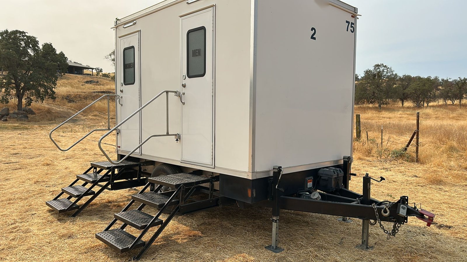 A Lavatory 2-Station restroom trailer on leveling jacks in a dry grass field with oak trees and a ranch fence