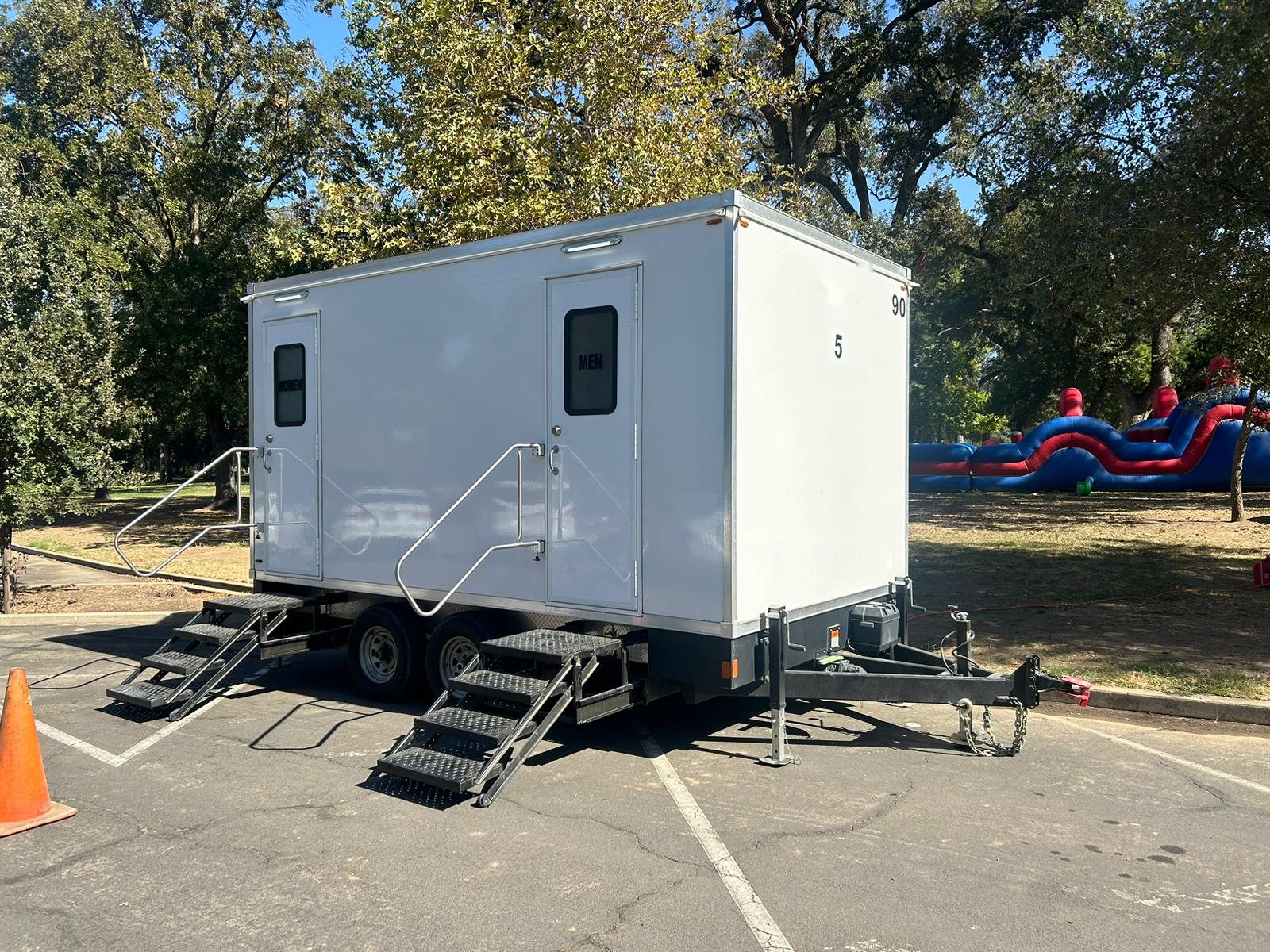 A Lavatory 5-Station restroom trailer in a park parking lot under shade trees, with an inflatable obstacle course on the lawn behind it