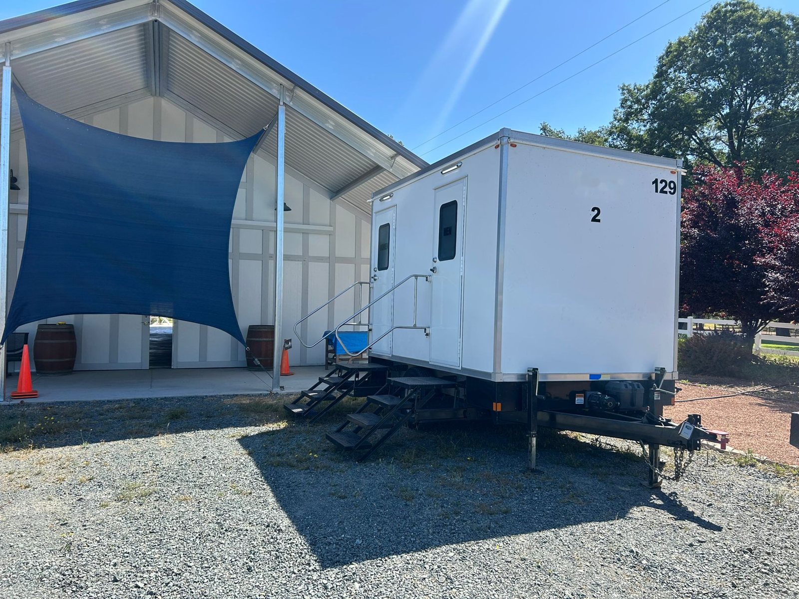 A Lavatory 2-Station restroom trailer on gravel beside a white barn venue with wine barrels and a shade sail