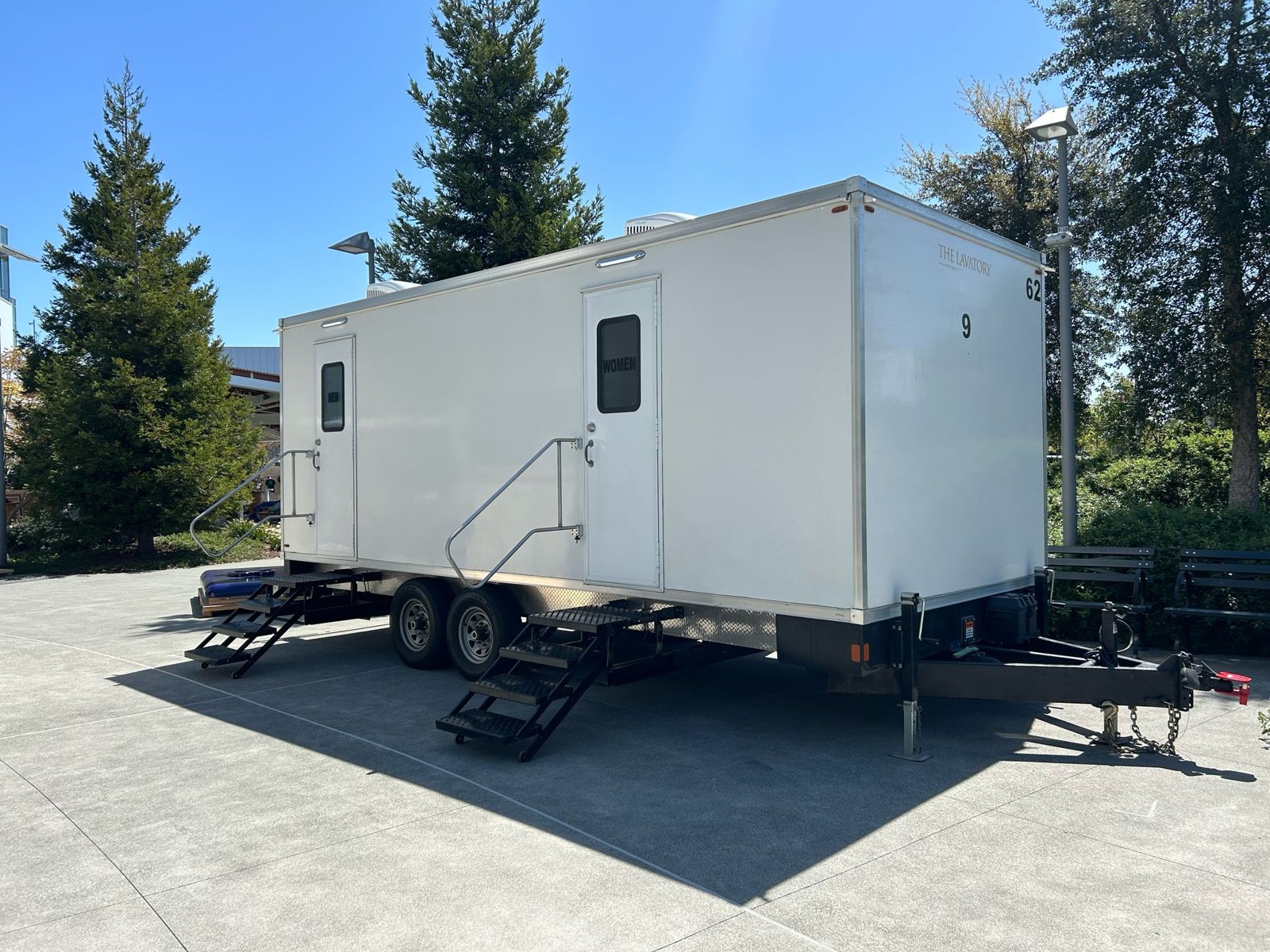 A Lavatory 9-Station luxury restroom trailer with steel stairs set on a concrete plaza between tall evergreen trees