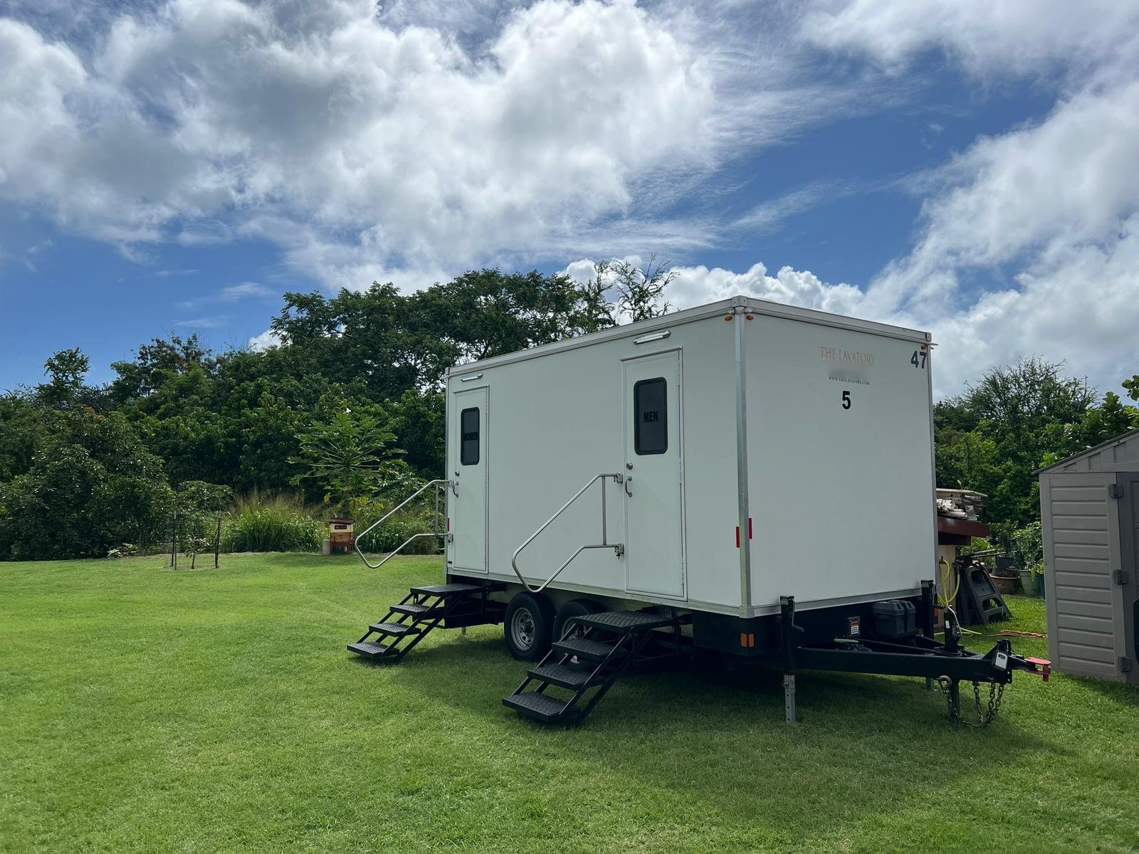 A Lavatory 5-Station restroom trailer set up on a green lawn in a backyard with trees behind it