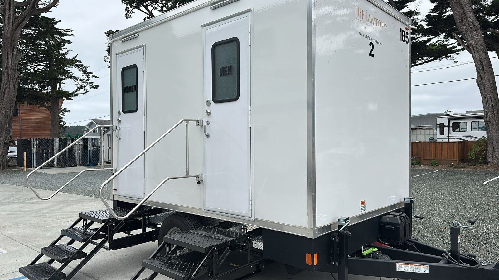 A Lavatory 2-Station restroom trailer with steel stairs and handrails parked on a gravel lot under cypress trees