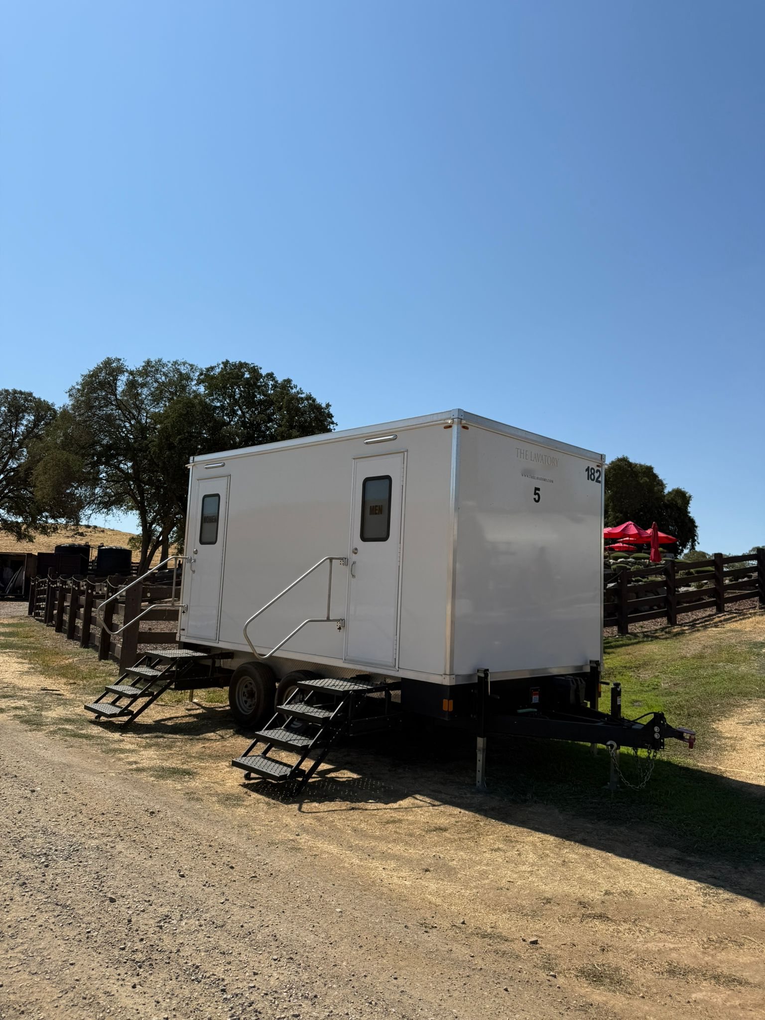 A Lavatory 5-Station restroom trailer on a dirt drive beside a wood rail fence, oak trees and red patio umbrellas
