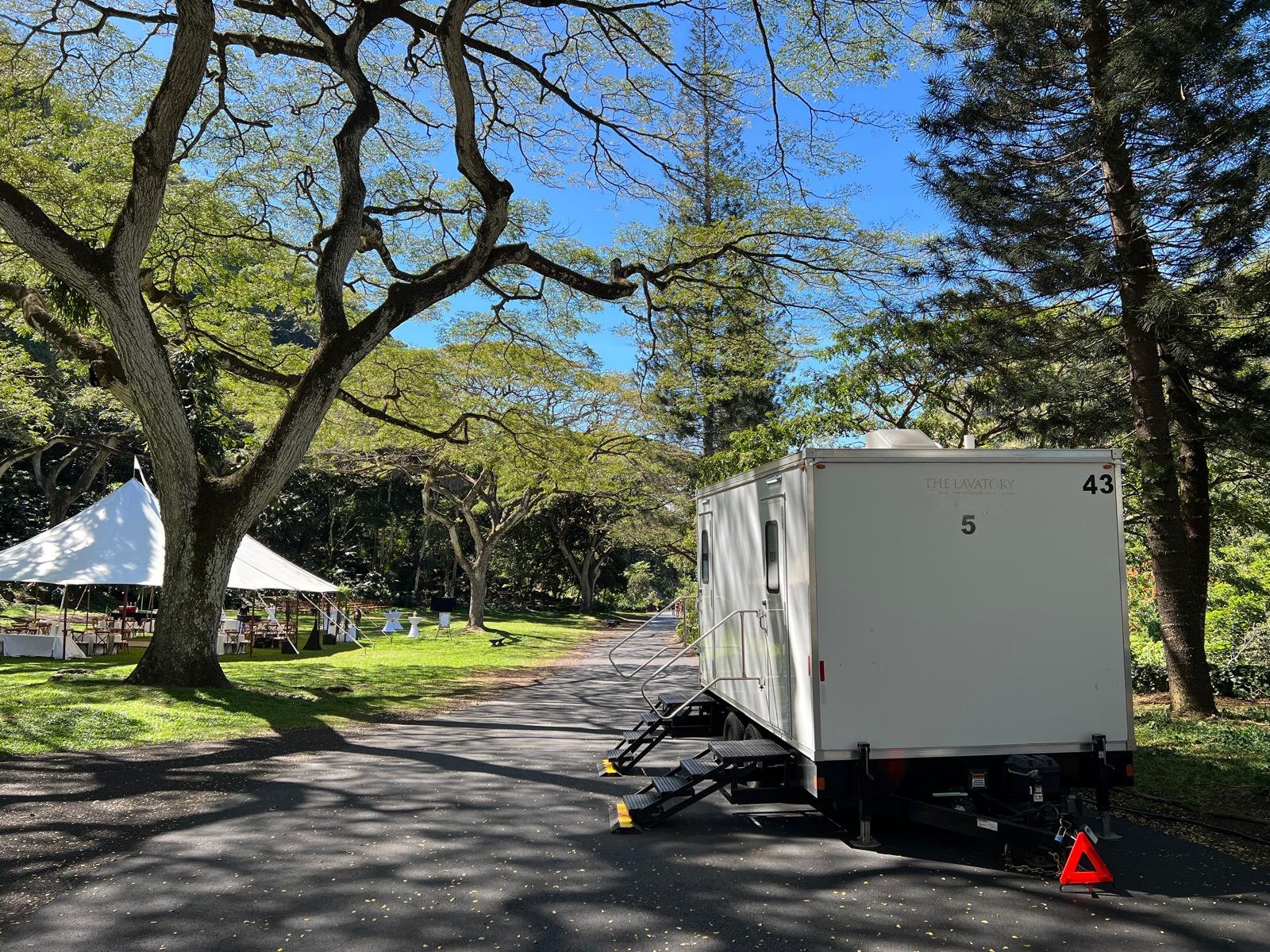 A Lavatory 5-Station restroom trailer on a shaded drive beside a white tent with reception tables under large trees