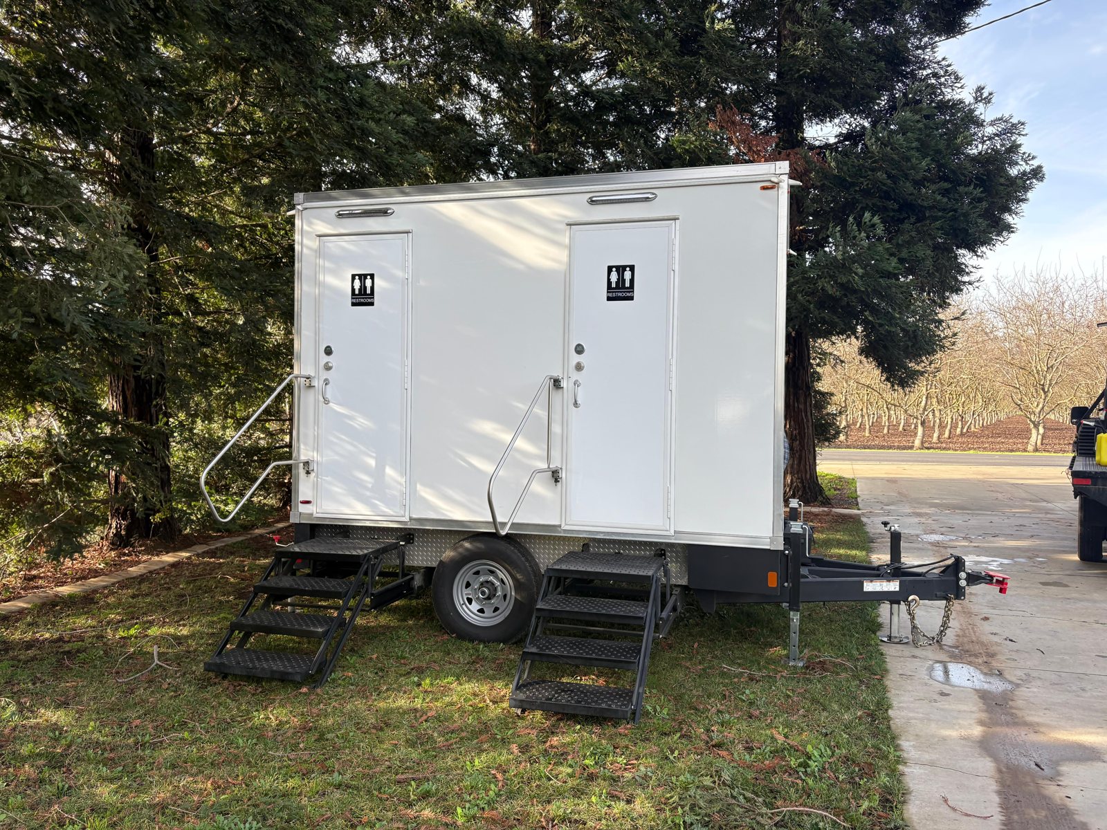 A Lavatory 2-Station restroom trailer on grass under tall evergreen trees, with a bare walnut orchard beyond the drive