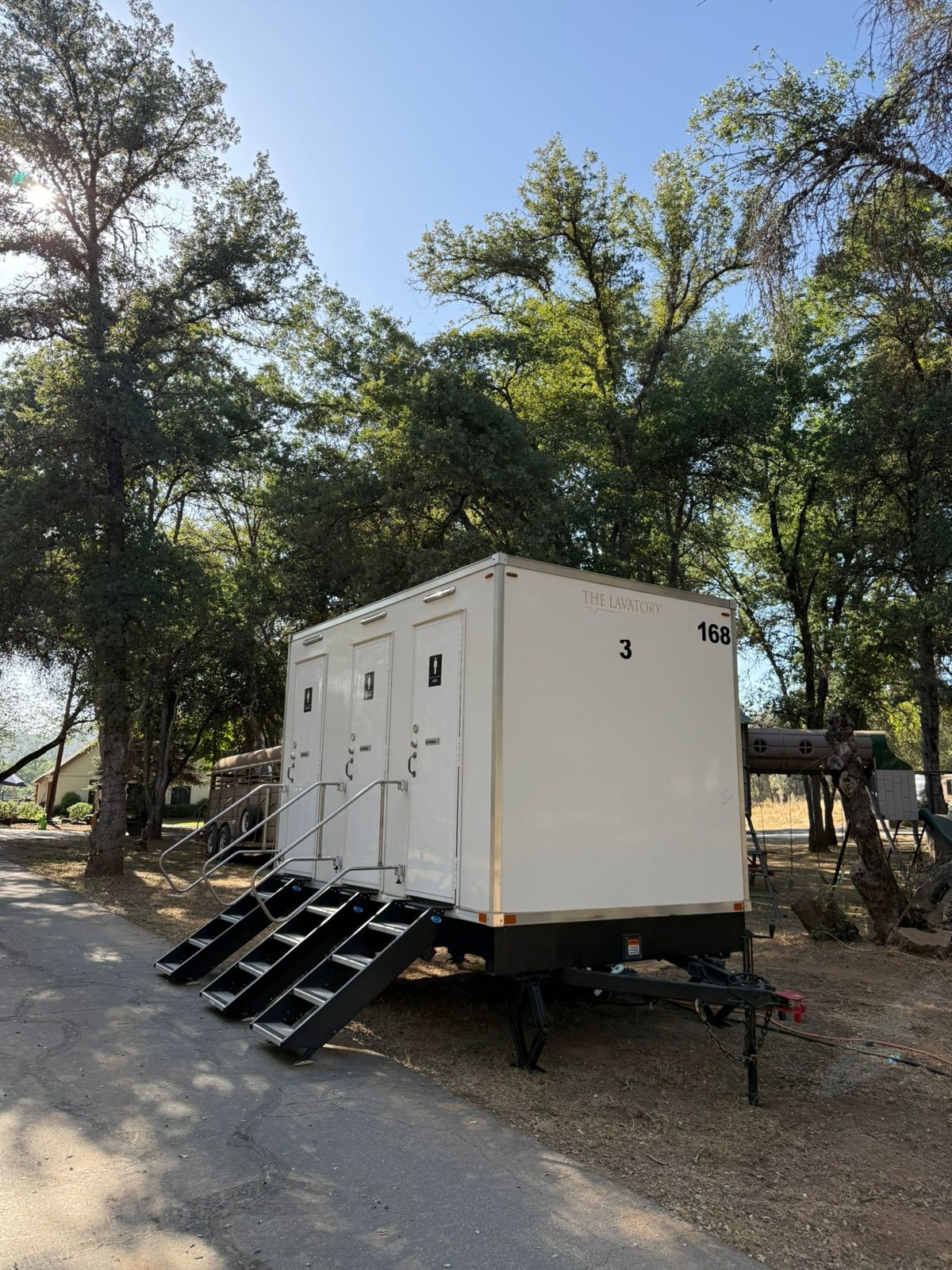 A Lavatory 3-Station restroom trailer set up under shade trees beside a paved path at a wedding site