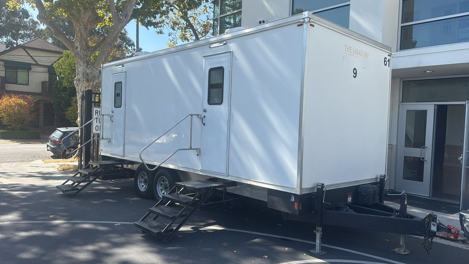 A Lavatory 9-Station restroom trailer parked on the pavement beside a modern office building entrance