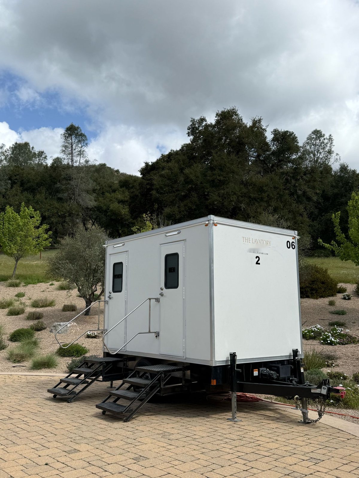 A Lavatory 2-Station restroom trailer with women's and men's doors on a paver patio in a garden with olive trees and oaks