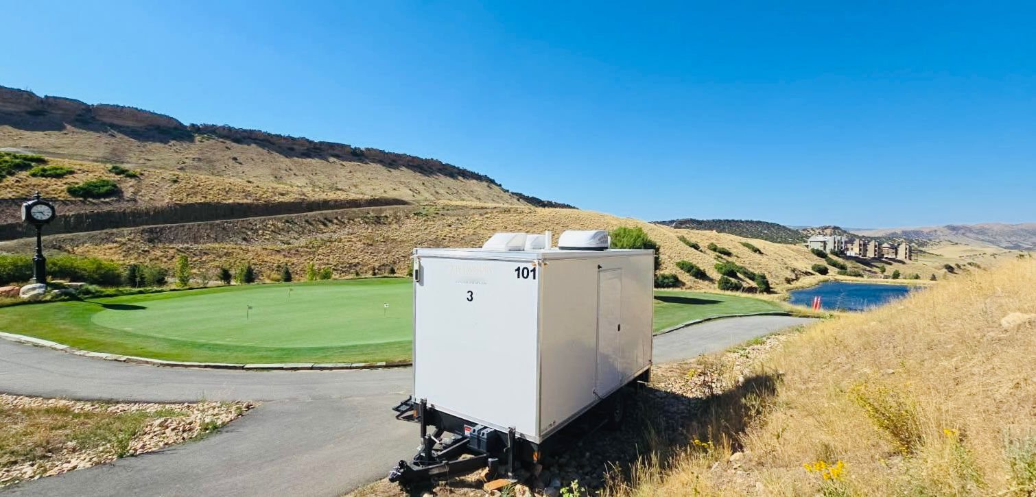 A Lavatory restroom trailer parked on a cart path beside a putting green, with dry foothills, a rock rim and a pond behind it
