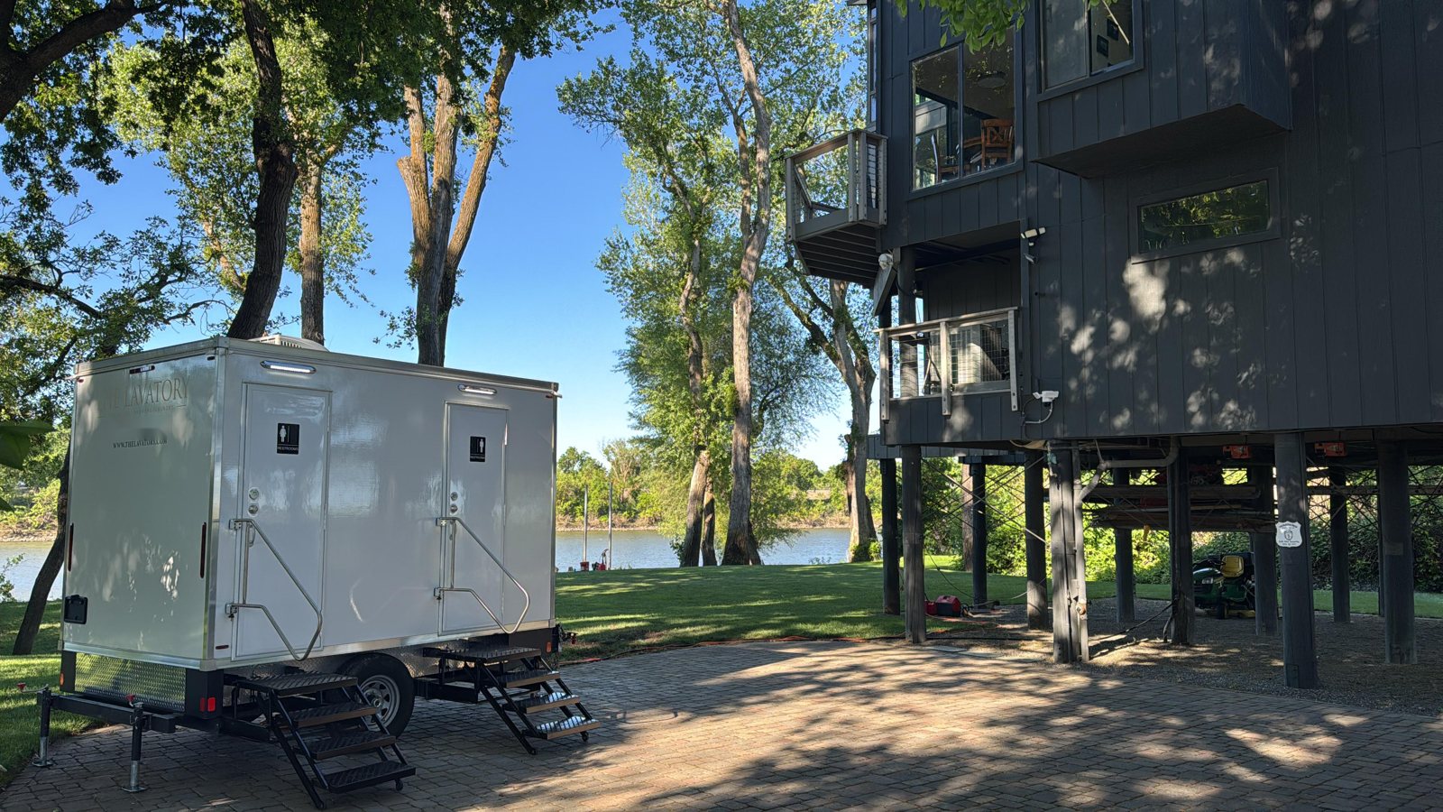 A Lavatory 2-Station restroom trailer on a paver drive beside a modern home on stilts, with tall cottonwoods and water behind it