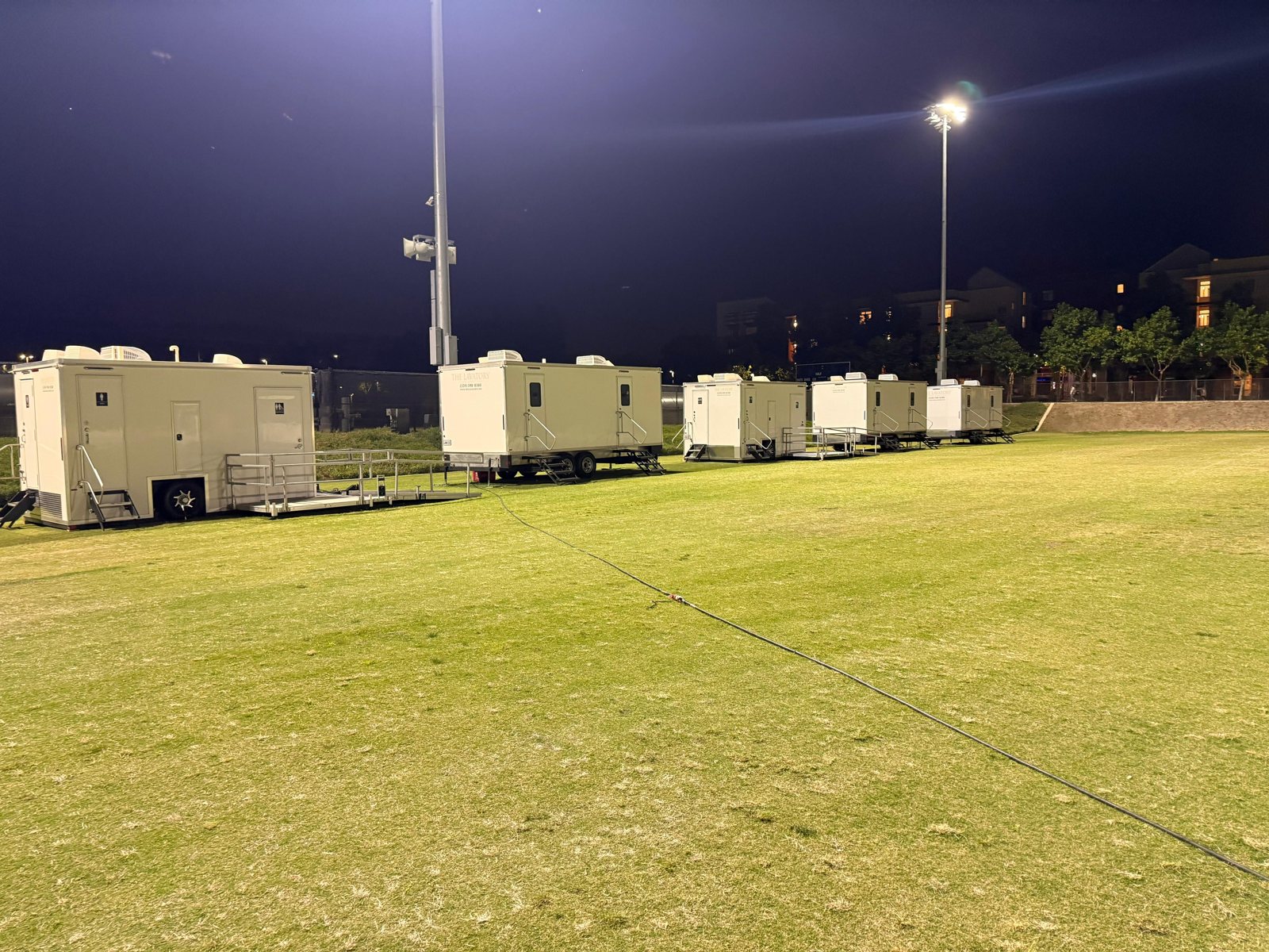 A row of Lavatory restroom trailers, including an ADA unit with a ramp, lined up along a lit sports field at night