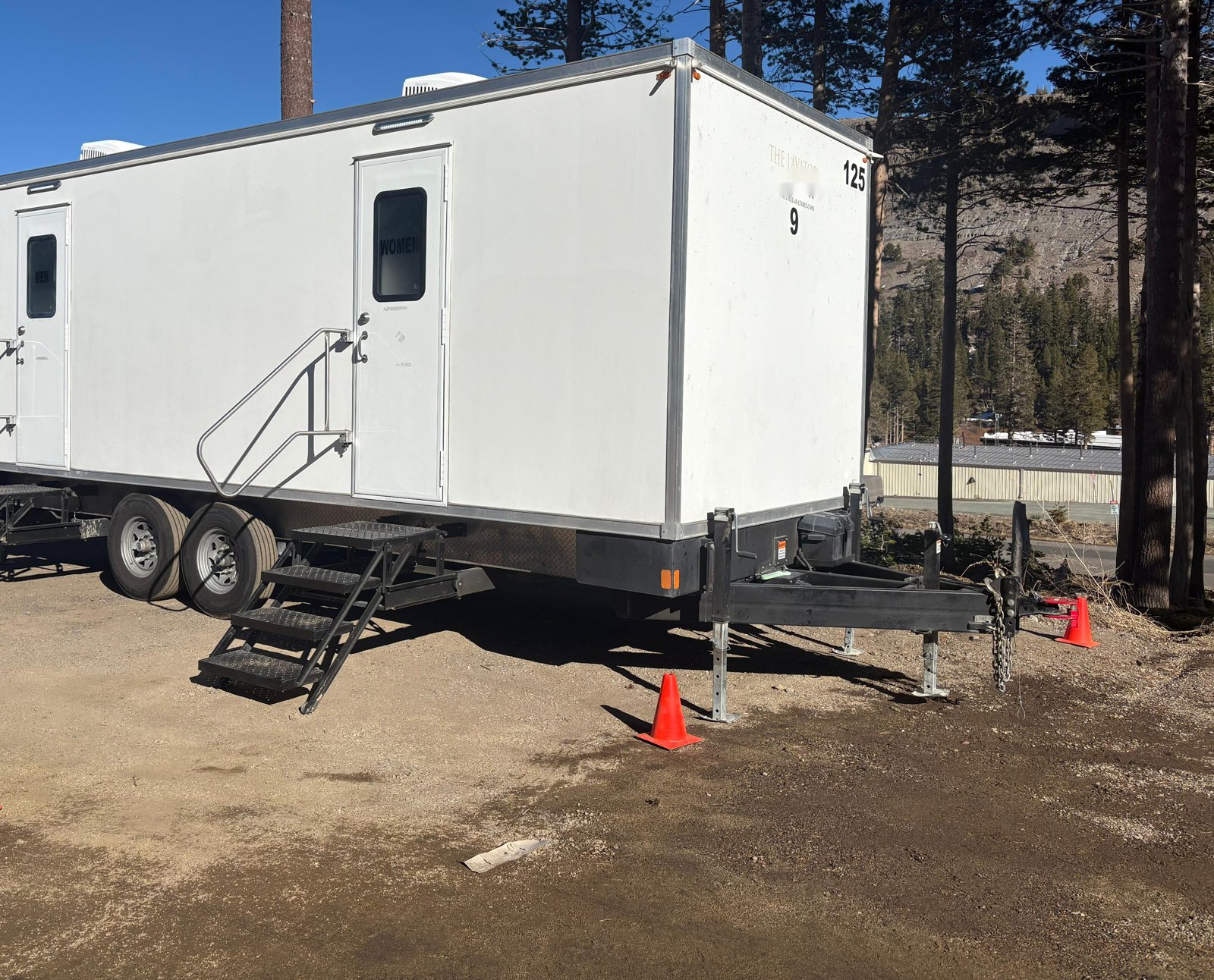 A Lavatory 9-Station restroom trailer with steel stairs at the women's and men's doors, parked on a dirt lot among tall pines below a rocky mountain slope