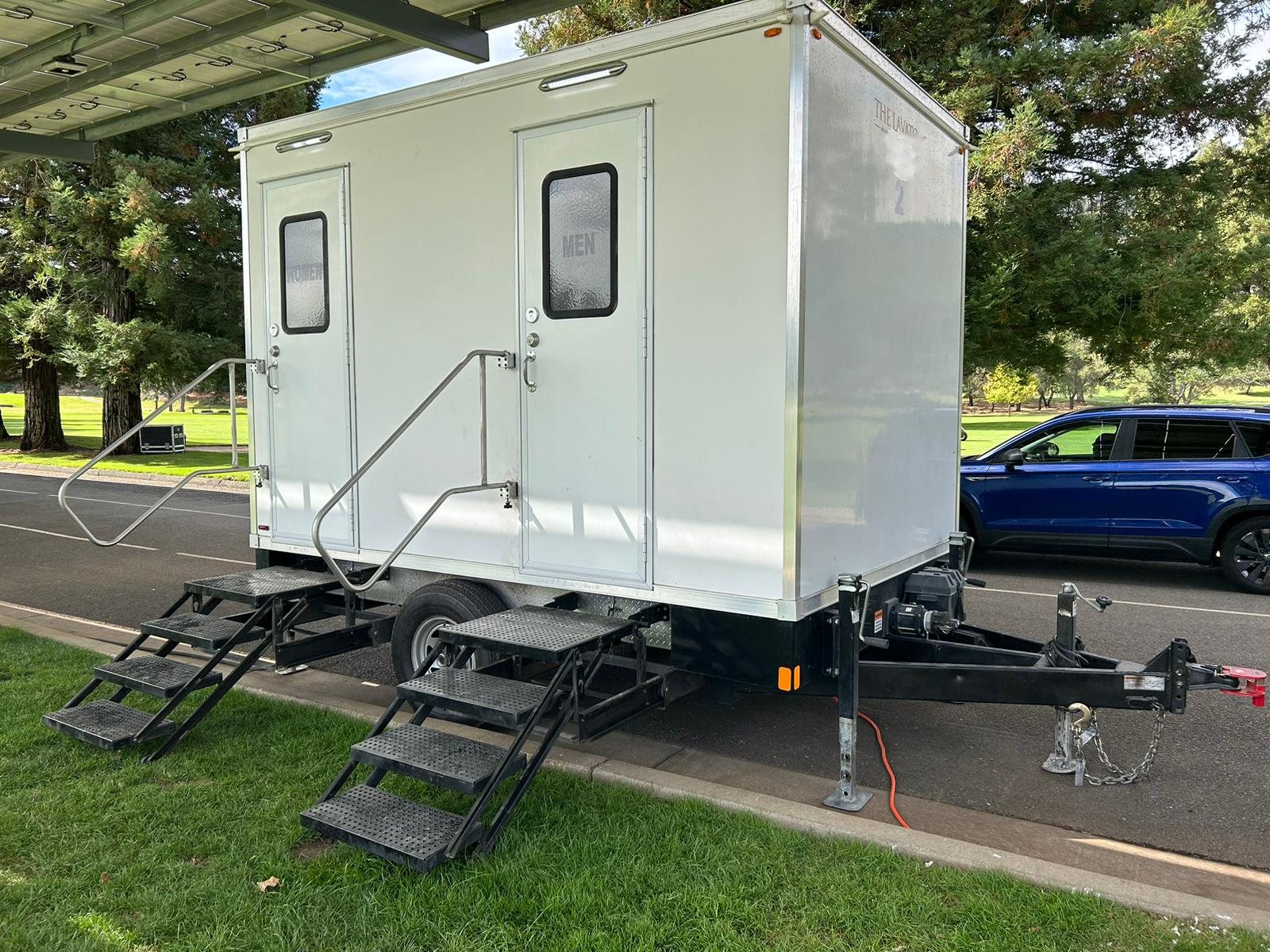 A Lavatory 2-Station restroom trailer parked at the curb beside a golf course lawn under tall evergreen trees, with steel stairs set on the grass