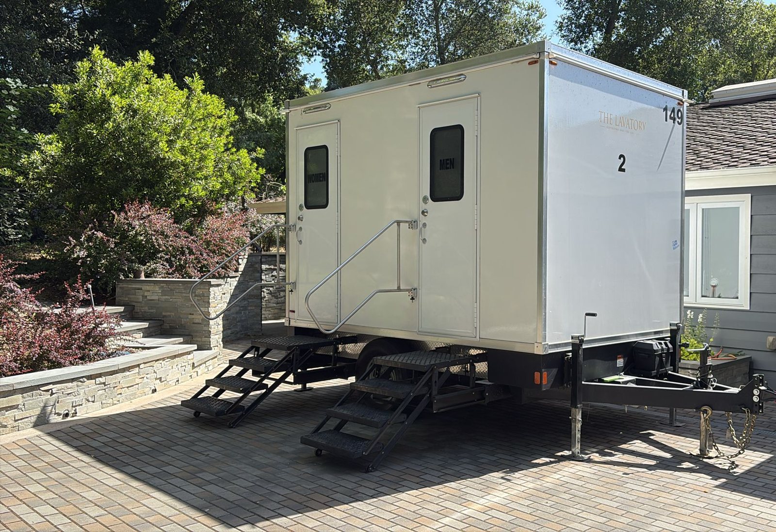 A Lavatory 2-Station restroom trailer parked on a paver driveway next to a gray house and a stone garden wall