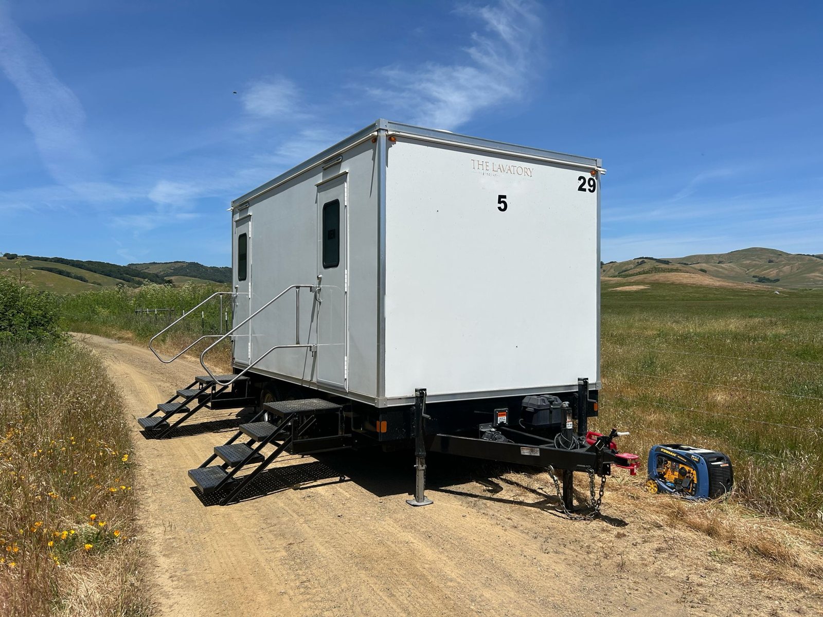 A Lavatory 5-Station restroom trailer set on a dirt ranch road between open grass pastures, with a small portable generator beside the hitch