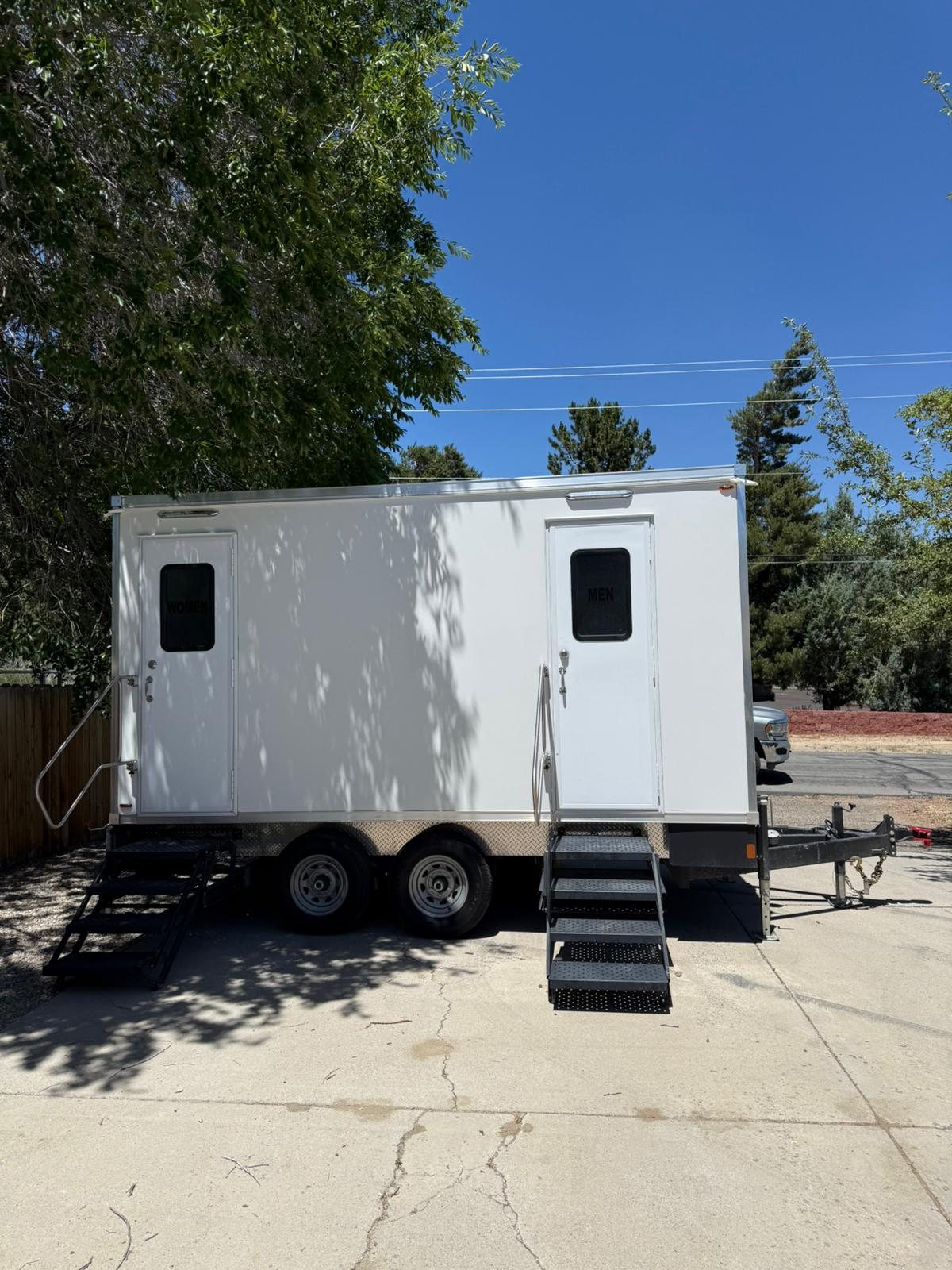 A Lavatory 2-Station restroom trailer with women's and men's doors and steel stairs on a home driveway under a shade tree