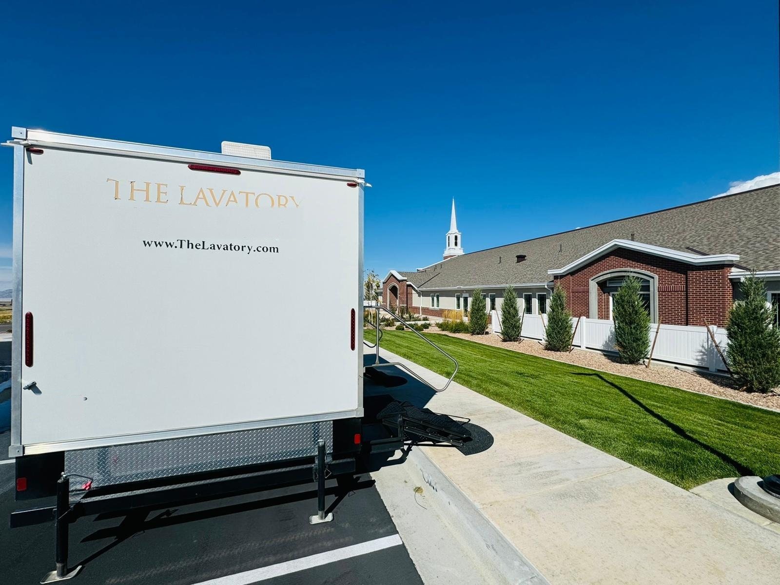 The rear of a Lavatory restroom trailer parked at the curb of a church parking lot, with a white steeple and a green lawn beside a brick building