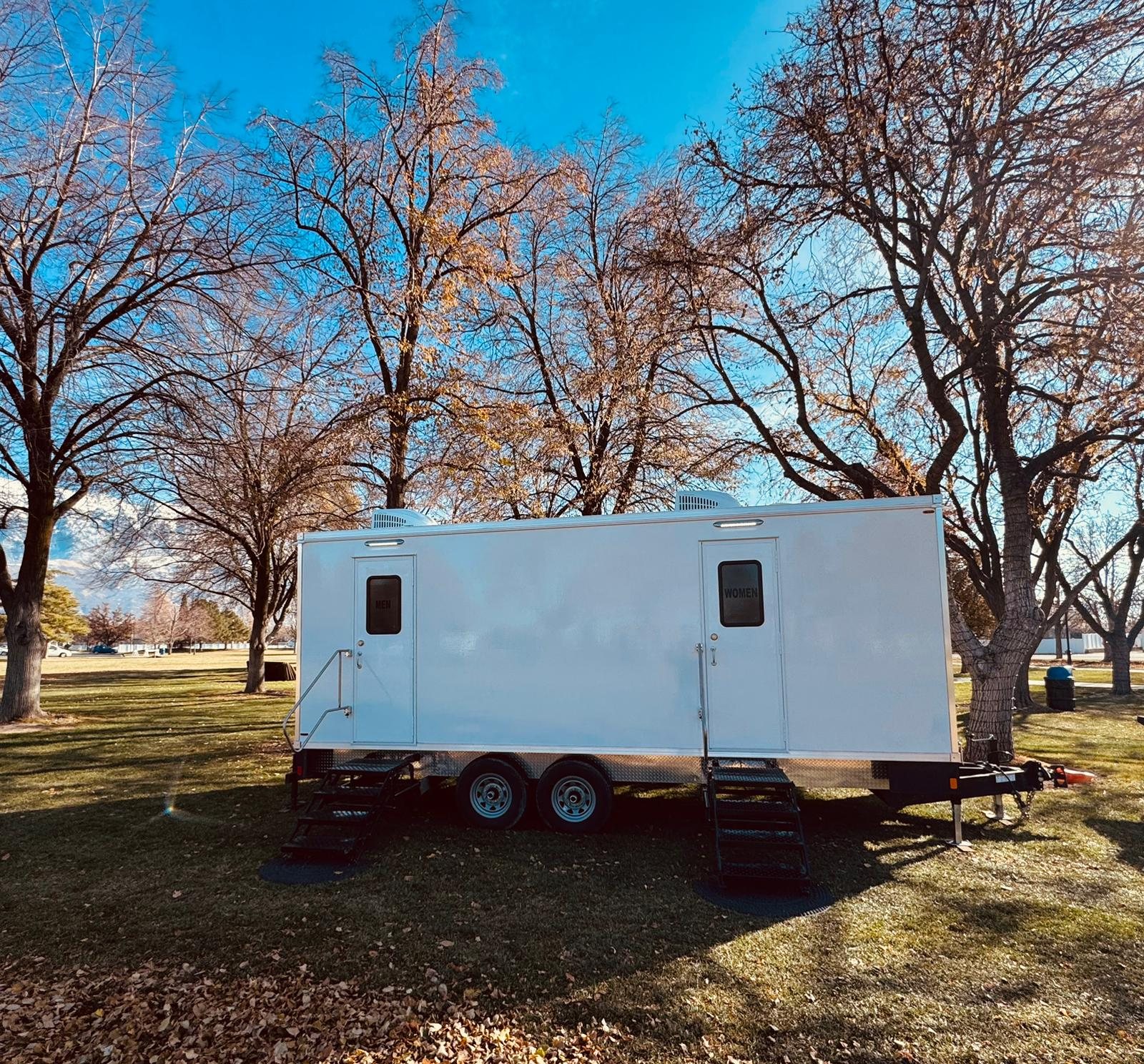 A Lavatory restroom trailer with separate men's and women's doors set on park grass under bare autumn trees