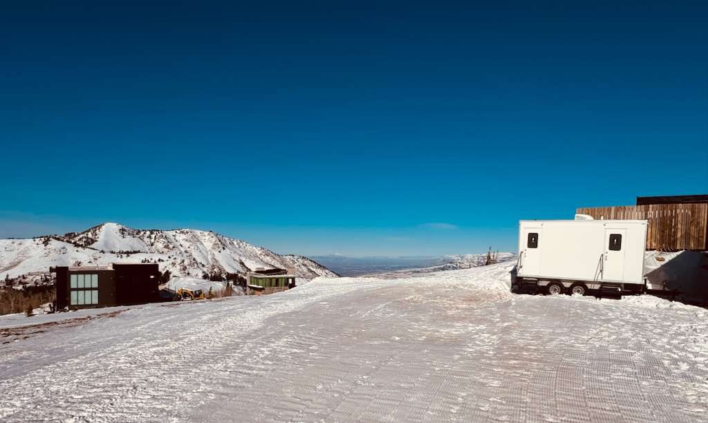 A Lavatory restroom trailer parked on a groomed snowy lot high in the mountains, with snow-covered peaks and a valley in the distance